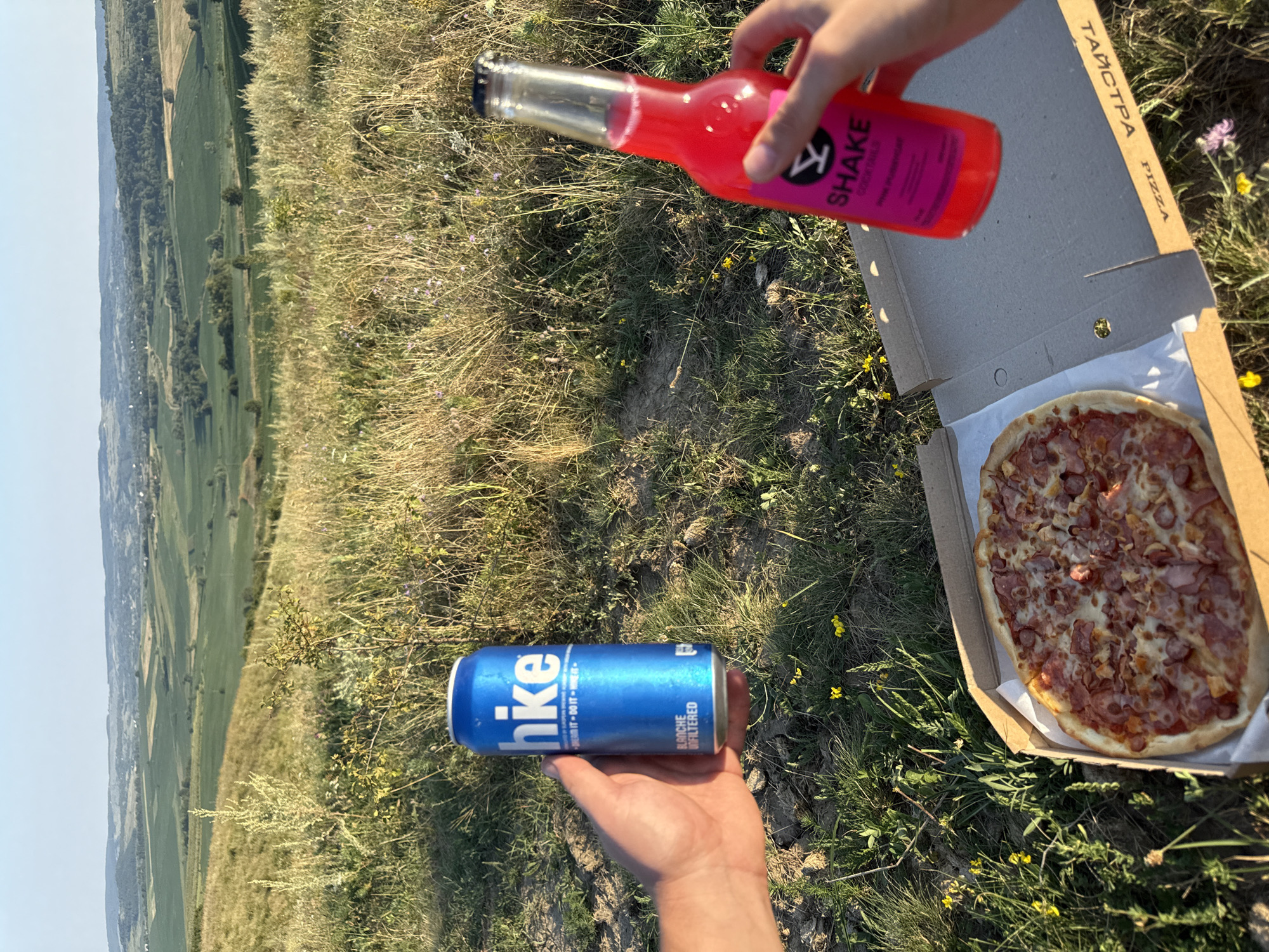 The image depicts a close-up view of a person's hands holding a blue aluminum can and a clear glass bottle filled with a pink liquid. In the foreground, a pizza in a cardboard box rests on the ground amidst dry grass and scattered yellow wildflowers. The background reveals a rolling landscape of green fields and trees under a clear blue sky. The can has "hike" written in white on it, and the bottle has a pink label with "SHAKE" and a stylized logo. The pizza appears to be topped with cheese and various meats, including what looks like sliced sausage and ham. The setting suggests an outdoor picnic or meal in a natural, rural environment.