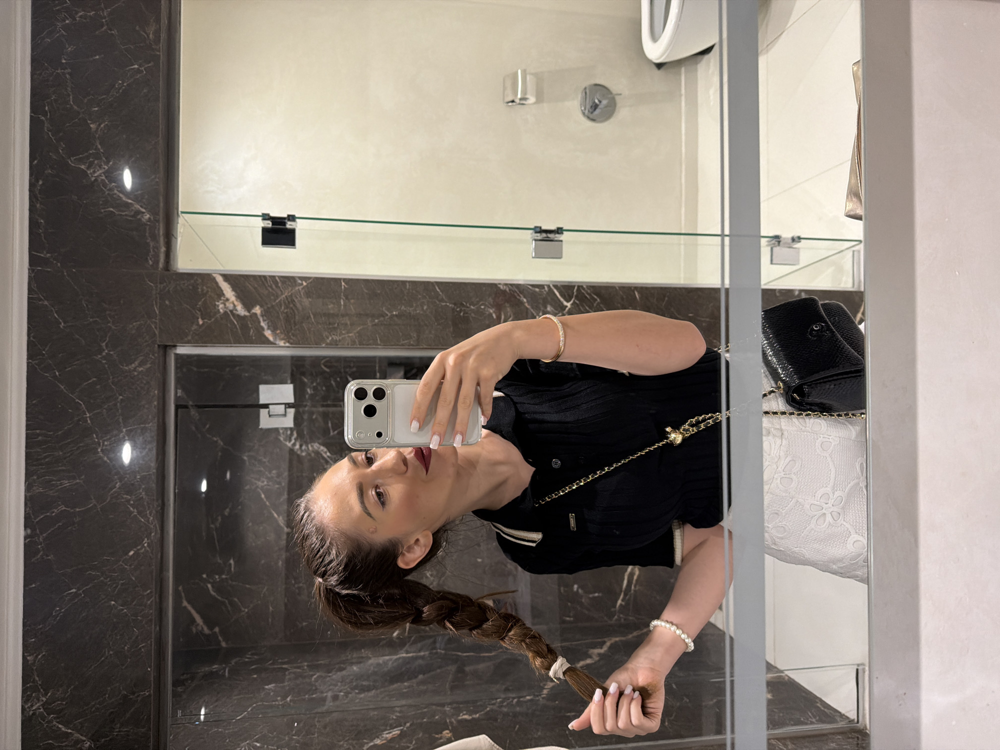 This is a mirrored selfie taken in a bathroom. The subject is a woman with long, braided brown hair, wearing a black top with a white collar and trim, and a pearl bracelet on her left wrist. She is holding a smartphone in her right hand, which is capturing the reflection. In the reflection, a black handbag with a gold chain strap is visible. The bathroom features dark marble walls and a large mirror. The woman appears to be taking a photo of herself while looking at her reflection. The overall mood is casual and personal.