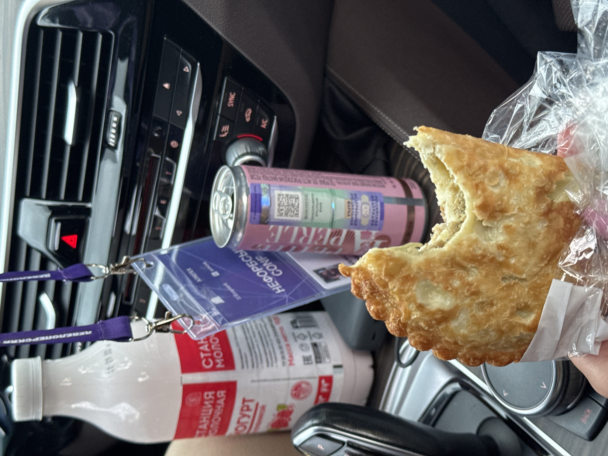 The image shows a close-up view of a person's hand holding a partially eaten savory pastry inside a plastic wrapper. The pastry is golden brown and appears to be flaky. In the background, several items are visible within a car's interior. To the left, a white bottle with a red label, likely containing yogurt or a dairy drink, is present. A purple lanyard with a clipped identification card hangs from above, partially obscuring the bottle. In the center background, a pink and blue can is situated next to the ID card. The car's dashboard, including air vents and control buttons, is visible. The location is identified as Estosadok, Russia, suggesting the scene takes place within a vehicle in this resort area.
