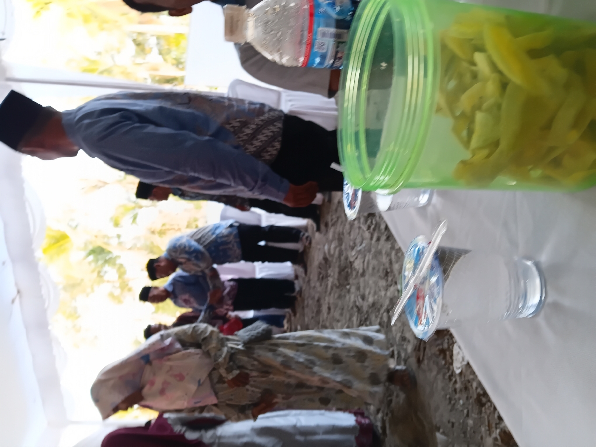 This is a medium-shot, slightly blurred, outdoor photograph taken in Sumber Mulyo, Indonesia. The scene appears to be a gathering or event taking place under a white tent. In the foreground, a white tablecloth covers a table. On the table, there are two clear plastic cups, one with a foil lid and a plastic straw, and a large green plastic container filled with sliced green fruit, possibly pickles or a type of fruit salad. Behind the table, several individuals are visible. On the left, a woman in a patterned headscarf and dress is walking away from the camera. To her right, a group of men are conversing. One man in the center is wearing a light blue patterned shirt and a black cap. Another man to his right is wearing a blue patterned shirt. Further back, more people are obscured by the tent and the blur. The ground in the mid-ground appears to be dirt or sand. The background shows foliage and trees, indicating an outdoor setting.