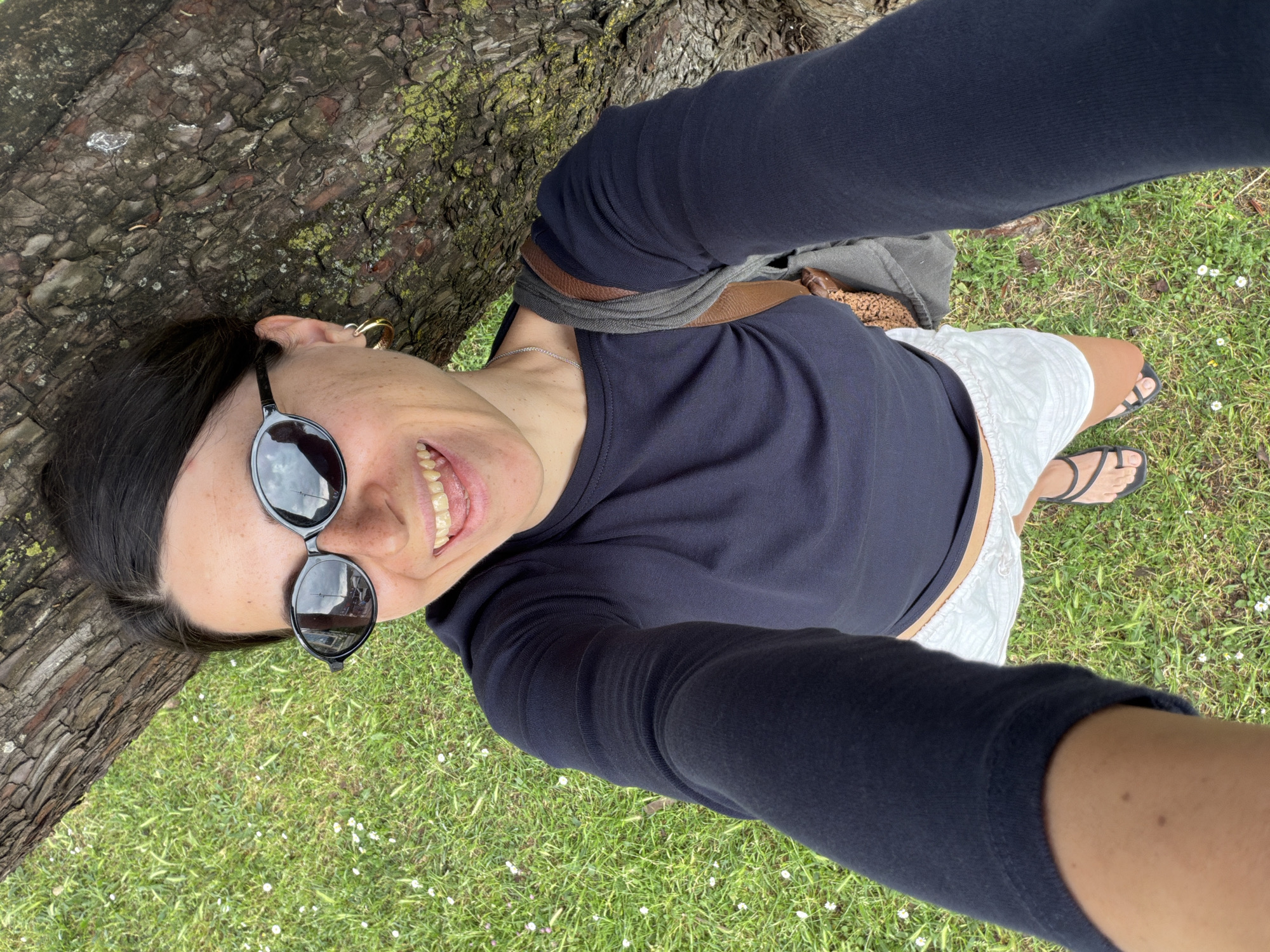 A woman is taking a selfie while leaning against a tree. She is wearing a dark long-sleeved shirt, white shorts, and black sandals. She has sunglasses on and is smiling. The background shows grass with small white flowers and the bark of the tree. The location is suspected to be Budva, Montenegro.