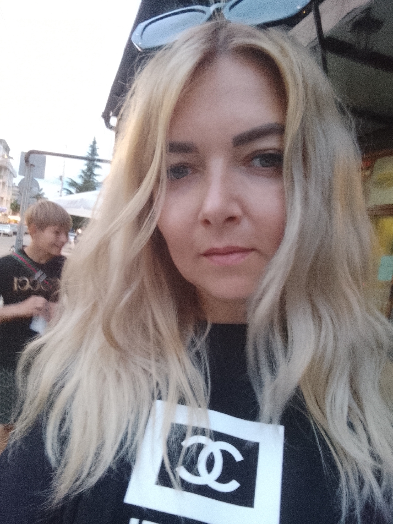 A blonde woman with a light complexion and blue eyes is taking a selfie outdoors. She is wearing a black t-shirt with a white Chanel logo. Her hair is long and wavy, and she has sunglasses on top of her head. In the background, a younger person with short brown hair is visible, wearing a black t-shirt with a red and green stripe across the chest. The setting appears to be a street in Nesebar, Bulgaria, with buildings and trees visible in the distance. The lighting suggests it is daytime, possibly late afternoon or early evening.