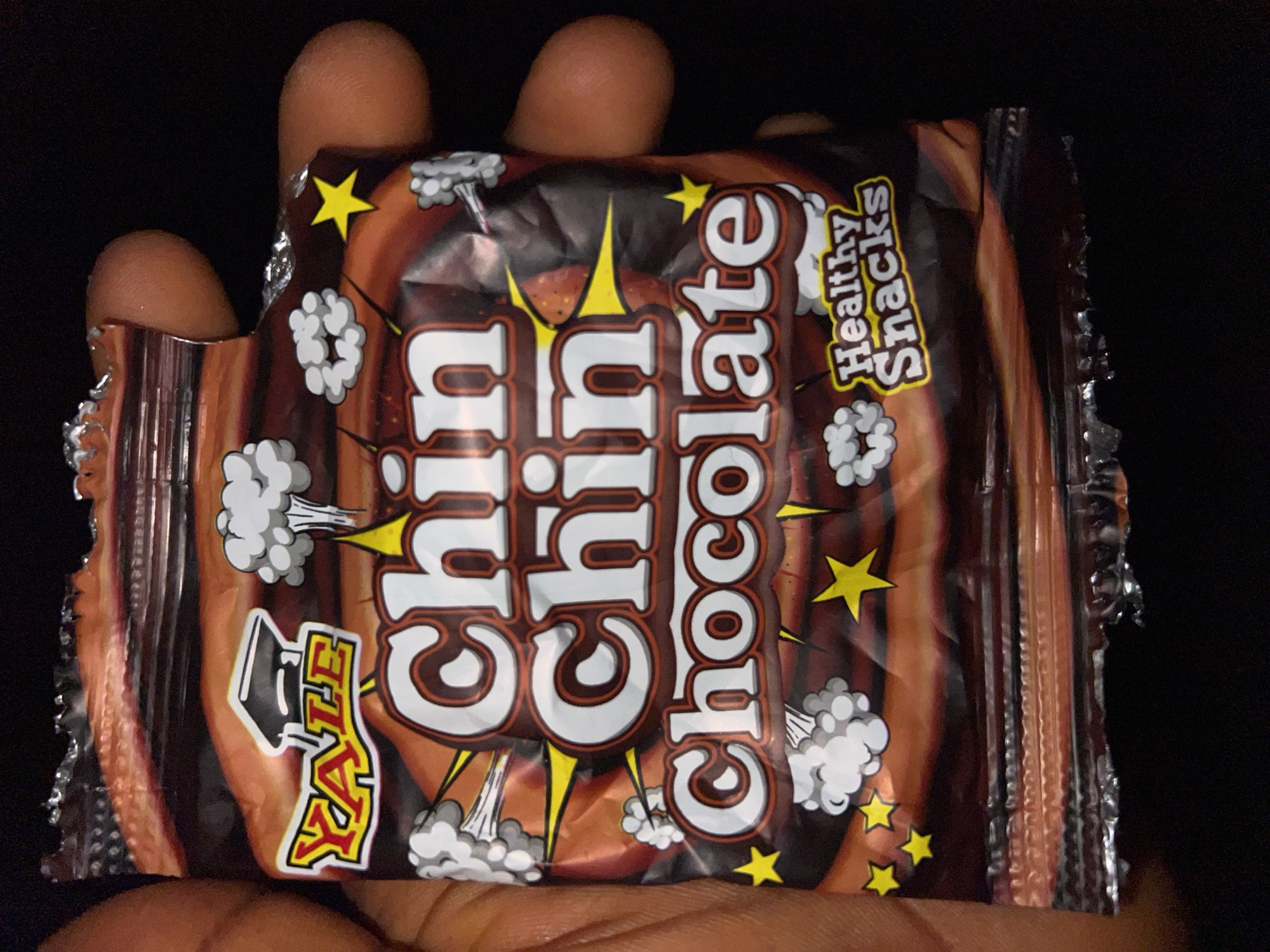 The image shows a close-up of a bag of "Chin Chin Chocolate" snacks being held by a person's hand against a dark background. The bag is primarily brown with text and graphics in white, yellow, and red. Prominently displayed are the words "Chin Chin Chocolate" in large, stylized white lettering with a dark outline. Above "Chin Chin", there is a yellow and red logo with the word "YALE" and a graduation cap graphic. To the sides of the text are cartoonish white cloudburst graphics with yellow accents. In the lower right corner, the words "Healthy Snacks" are written in white with yellow outlines on a yellow background. The bag appears to be made of a shiny material, and its bottom edge is torn and crimped. The person's fingers are visible on the left and right sides of the bag, indicating the snack is being held. There is no identifiable context of Lagos, Nigeria within the image itself.