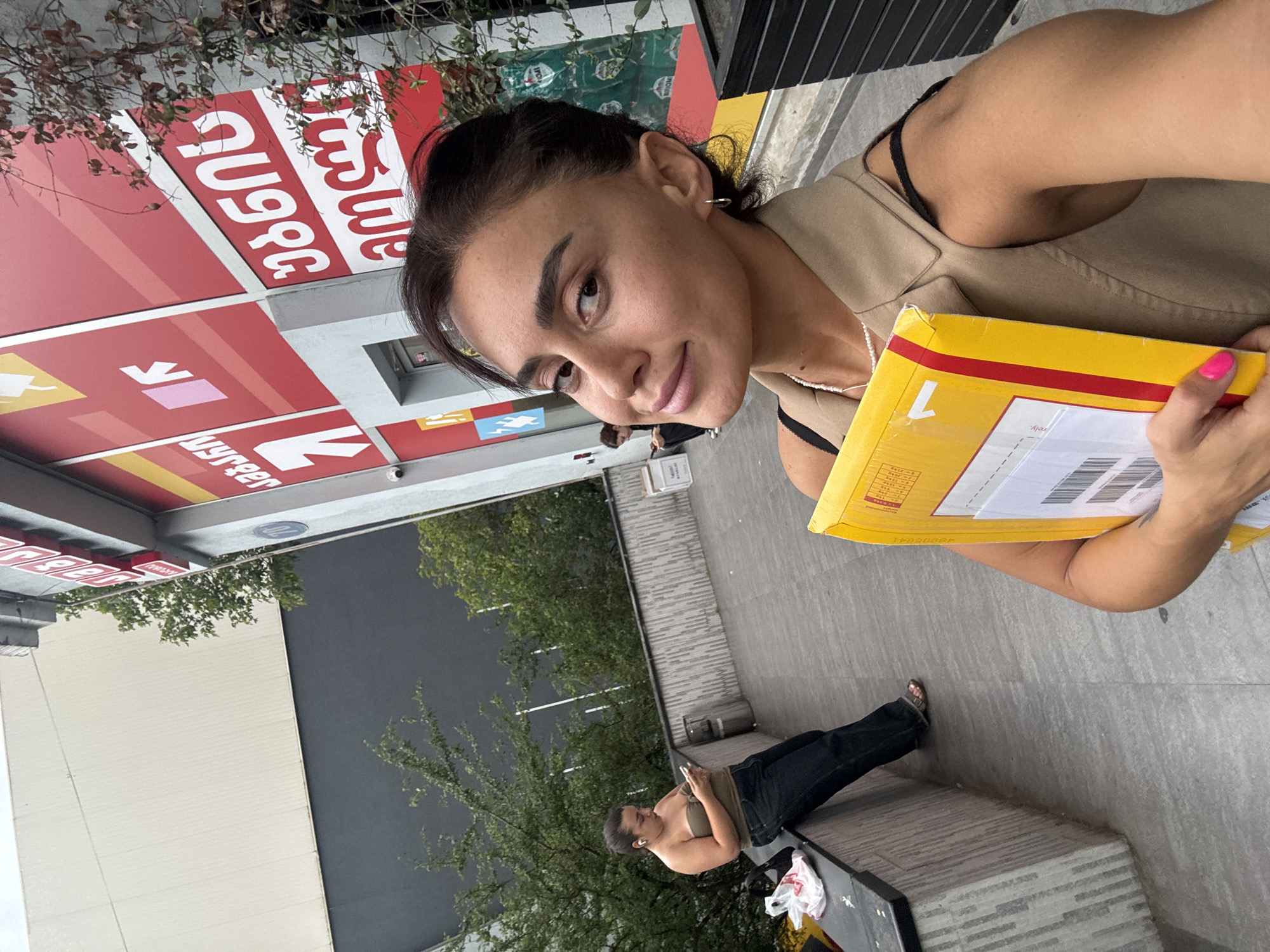 The media file shows a woman taking a selfie outdoors, holding a yellow package. She is wearing a tan vest over a black top, and has dark hair pulled back. Her nails are painted bright pink. In the background, there is a building with red signage featuring white text in Georgian script. Another person is visible sitting on a bench in the background, looking at their phone. The scene appears to be a street or public area, possibly near a business given the signage. The lighting suggests it might be daytime. The overall mood seems casual and relaxed. The visible text on the package is "DHL" and a barcode. The red signage on the building has Georgian script that appears to read "ბანკი" (bank).