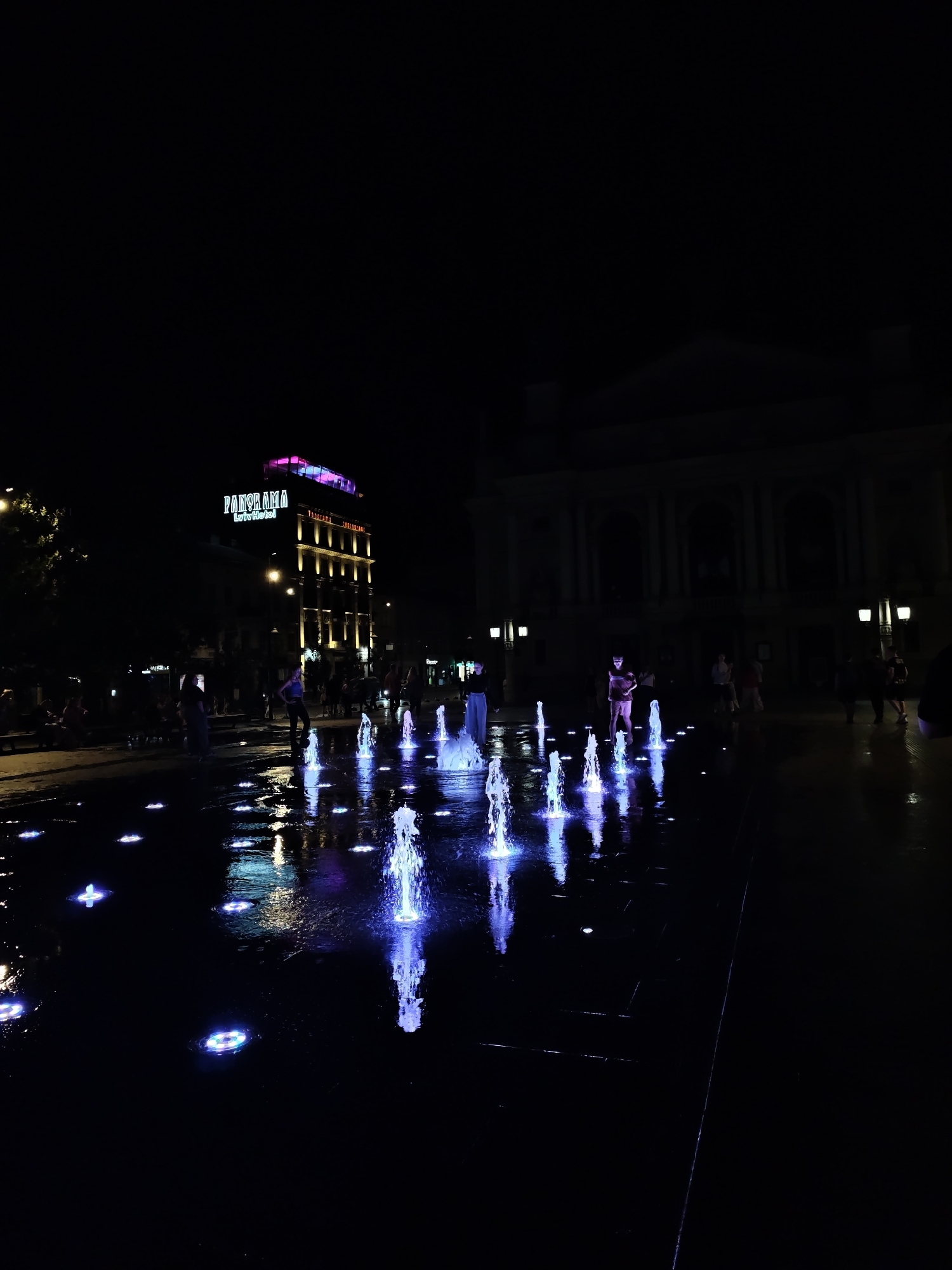 The image depicts a night scene at a modern water fountain plaza in Lviv, Ukraine, likely near the Panorama Lviv Hotel. The plaza features numerous ground-level water jets, some illuminated with vibrant blue and pink lights, creating a mesmerizing display on the wet pavement. Several people are present, scattered throughout the plaza. Some individuals are standing near or within the fountains, interacting with the water, while others are observing the spectacle or conversing. In the background, a multi-story building with illuminated windows and a rooftop sign that reads "PANORAMA Lviv Hotel" is visible. To the right, a neoclassical building with a grand facade and arched windows stands in silhouette against the dark sky, suggesting a historical or cultural institution. Streetlights illuminate the edges of the plaza, casting a soft glow on the surroundings. The overall atmosphere is one of a lively urban gathering space at night, characterized by the interplay of light, water, and human activity.