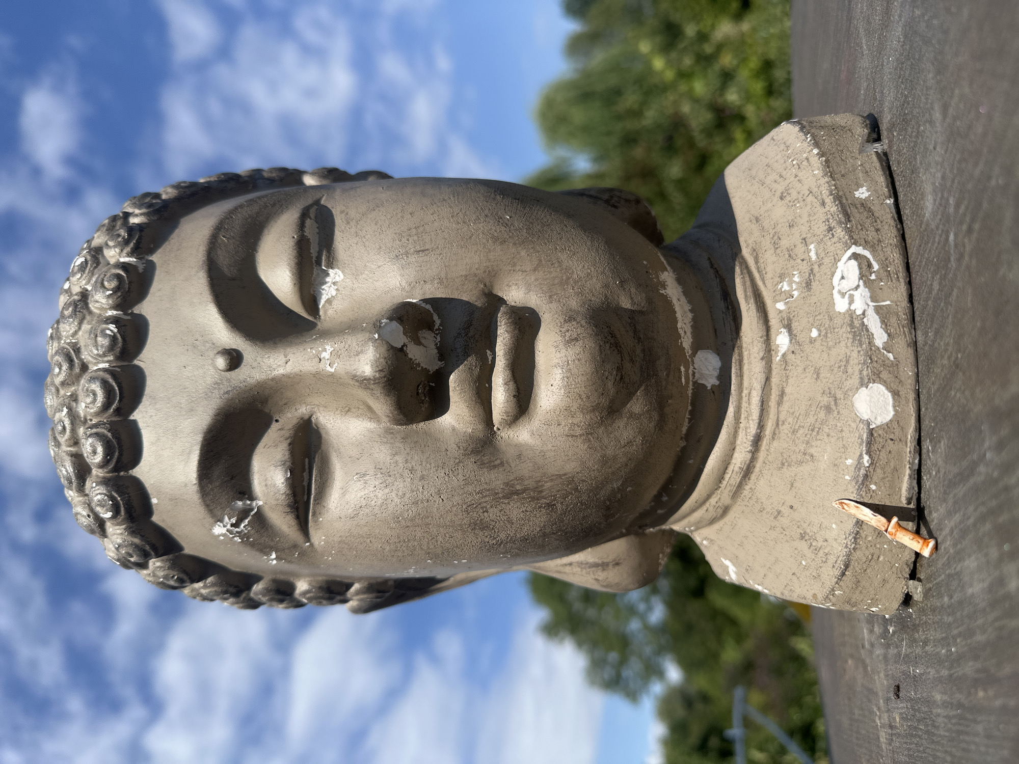 A close-up, eye-level outdoor shot features a weathered, stone statue of a Buddha's head. The statue, a neutral grey with chipped white paint accents, sits on a dark, textured surface, likely a wooden table or bench. A small, orange-handled toy knife is propped against the base of the statue on the right side. The background is blurred, showing green foliage and a bright blue sky with scattered white clouds. This scene is not located in Lelystad, Netherlands; it is an outdoor setting with no specific urban landmarks visible.
