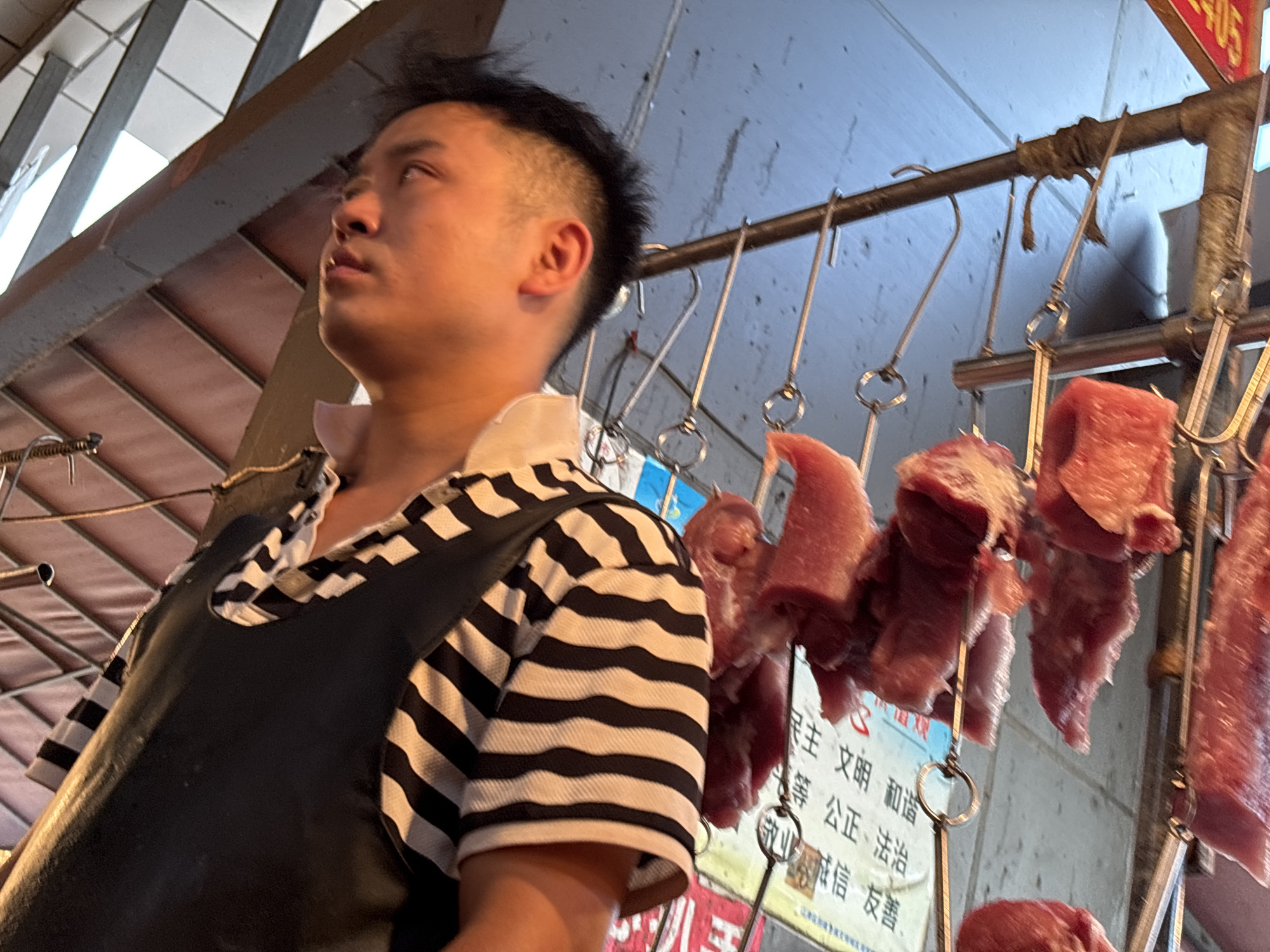 In a dimly lit outdoor market stall in Jiangjin, China, a young Asian man with a mohawk-style haircut is seen from the chest up. He wears a black apron over a black and white striped shirt. The apron is dark and appears to be made of a sturdy material, possibly leather or vinyl, and covers his torso. The striped shirt has thin black and white horizontal bands. His expression is neutral as he looks slightly upwards and to his left. Behind him, hanging from a metal rod by hooks, are several pieces of raw red meat. The meat is unevenly cut and appears to be fresh. The background consists of a rough, concrete-like wall and a ceiling structure made of exposed beams and reddish-brown panels, suggesting an open-air market setting. A poster with Chinese characters is partially visible behind the hanging meat, indicating a commercial establishment. The overall scene captures a moment in the daily operations of a butcher's stall.