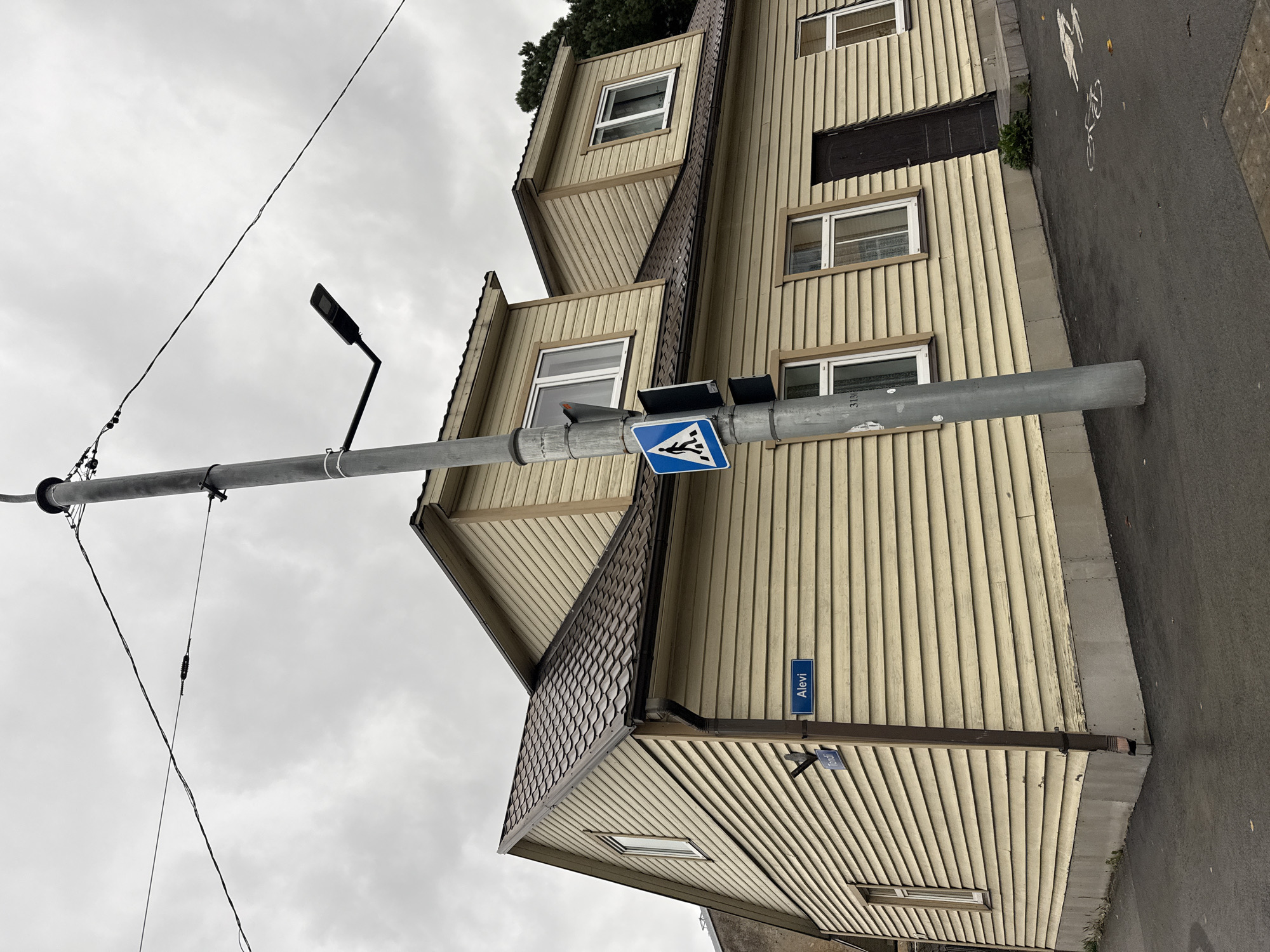 The image shows a street scene in Tallinn, Estonia, on a cloudy day. A light-colored wooden house with a dark tiled roof dominates the frame. The house has multiple windows and dormer windows. A street sign reading "Alevi" is mounted on the side of the house. A lamppost with a modern LED street light stands in front of the house, and a pedestrian crossing sign is attached to it. The street in front of the house is paved, and there are white markings on the pavement that appear to indicate a bicycle lane and a pedestrian crossing. The overall impression is that of a residential street in a European city.