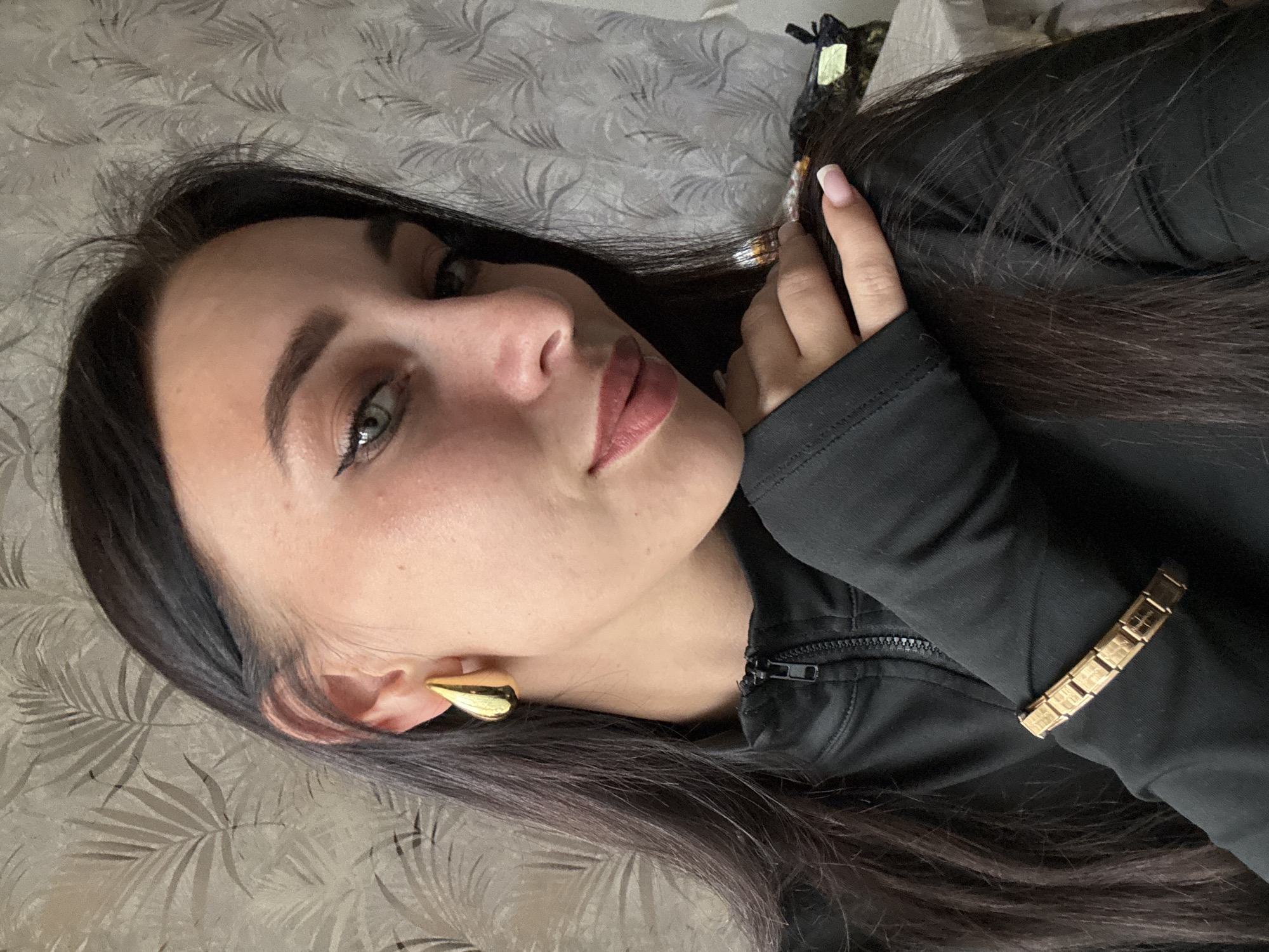 An indoor portrait shows a young woman, likely in her 20s or early 30s, looking directly at the camera with a calm expression, hinting at a subtle smile. She has long, dark brown hair, light-colored eyes (possibly green or hazel), and neatly groomed eyebrows. She appears to be wearing light makeup, including eyeliner and lipstick in a muted, rosy shade.
She is dressed in a black, long-sleeved top or jacket with a visible zipper detail on the front. On her left ear, she wears a prominent gold-colored earring shaped like a curved spike or teardrop. A gold-toned bracelet with rectangular, interconnected links, possibly featuring an indiscernible embossed pattern or text, adorns her right wrist. Her fingernails are manicured in a French style with pink bases and white tips. One hand is gently placed near her head, touching her hair, which is partially secured by a dark hair clip visible at the top.
The woman is positioned in a reclining or lying pose, suggesting the photo was taken from above. The background features wallpaper with a repeating, subtle gray pattern resembling leaves or ferns. The setting is an interior space in Chaikovsky, Russia. The lighting is even, suggesting ambient indoor light. No clear text is legible on the bracelet or elsewhere in the image.