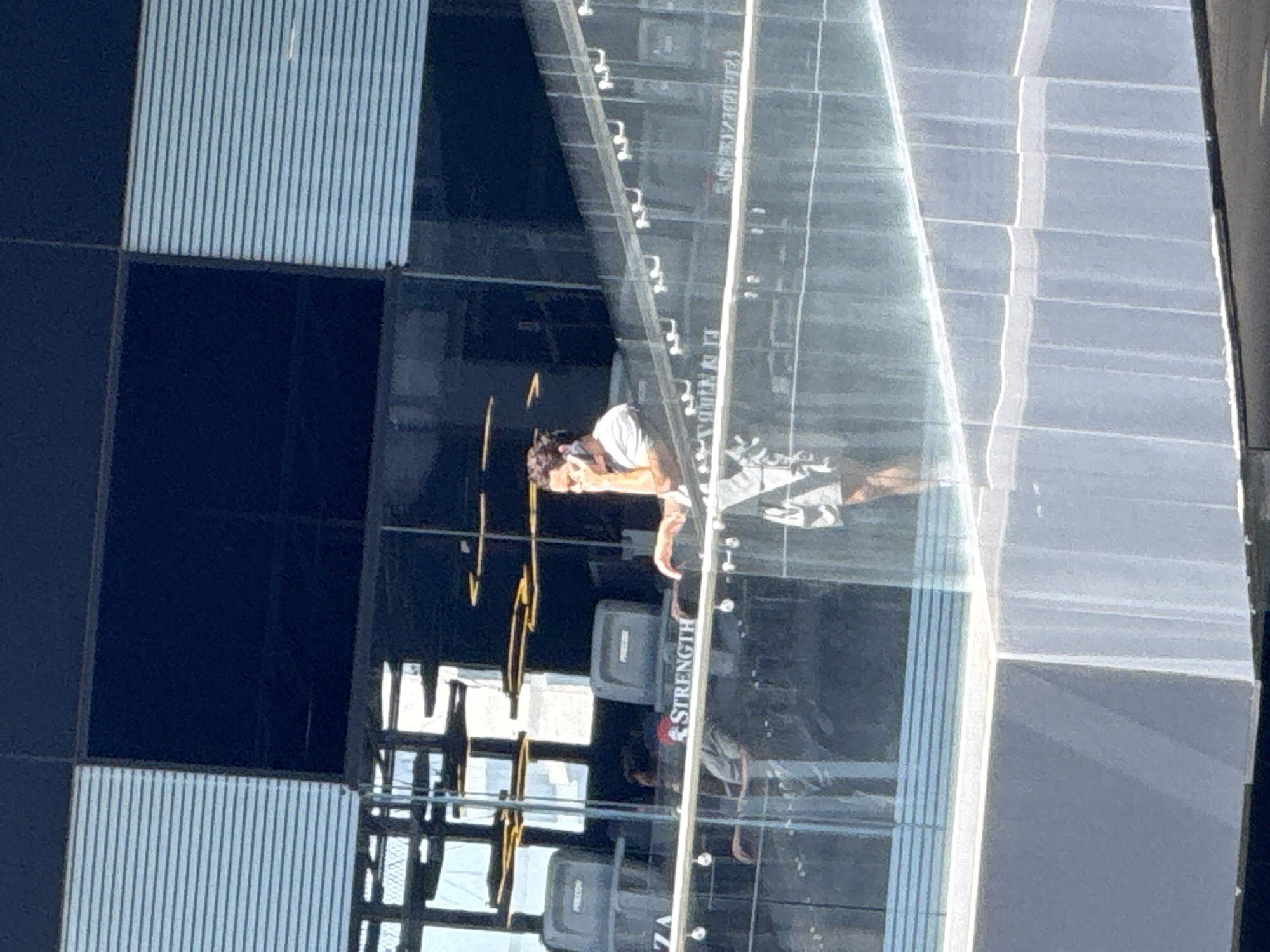 The image, taken in San Bernardino Tlaxcalancingo, Mexico, captures a brightly lit modern fitness facility, likely a gym, characterized by extensive glass architecture. The scene features a person with dark hair tied back, wearing a light-colored top, standing on the right side of the frame and looking towards the left.
The facility showcases large glass partitions that serve as walls, reflecting the interior elements and creating a bright, open atmosphere. Visible in the reflections are other individuals and parts of the building's structure. On the left side of the room, the walls are composed of alternating dark, possibly matte, panels and light-colored panels with fine vertical stripes.
Several pieces of gym equipment are present, including machines branded "PRECOR." The word "STRENGTH" is clearly visible, printed vertically on a panel, likely part of the fitness equipment or a decorative element. Abstract golden, linear structures are also visible in the mid-left area of the image, potentially serving as art installations or decorative lighting. The overall impression is one of a clean, contemporary fitness environment under clear daytime lighting.