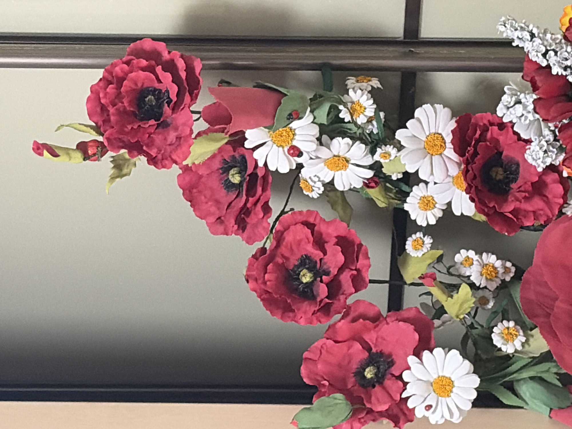 A close-up, eye-level shot showcases a vibrant artificial floral arrangement against a neutral background, likely an interior wall with a partition. The arrangement features several large, deep red poppies with dark centers and delicate, ruffled petals. Interspersed among the poppies are numerous white daisies with bright yellow centers. Two small ladybugs, one on a daisy petal and another near a daisy, add a whimsical touch. Green leaves and stems provide a naturalistic backdrop to the flowers. The overall impression is one of a cheerful and decorative display. The image does not contain any elements that would indicate its location in Chaikovsky, Russia.