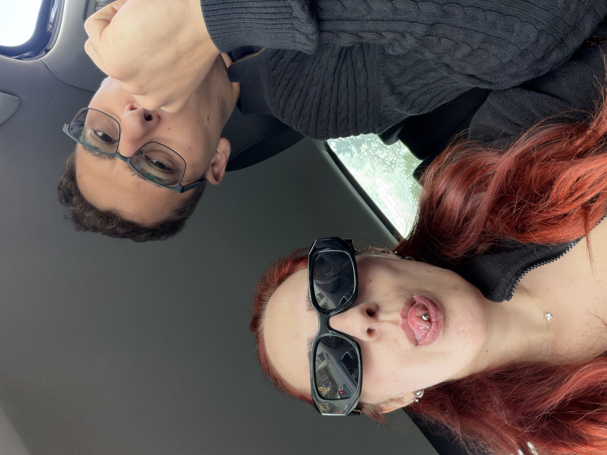 Two individuals are in a car. One person has brown hair, glasses, and is wearing a black sweater. The other has red hair, sunglasses, a pierced tongue and a star necklace. The picture is taken from above and is slightly rotated. The location is unknown.