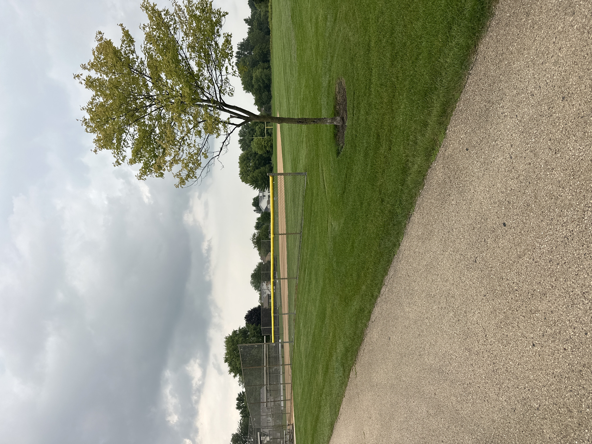 The image depicts an outdoor scene on an overcast day, likely in a park or recreational area. In the foreground, a paved pathway with a speckled beige texture runs diagonally from the lower left to the upper right of the frame. To the right of the pathway is a meticulously manicured lawn of vibrant green grass.
Beyond the lawn, a chain-link fence encloses a baseball field. The baseball field itself is visible with its reddish-brown dirt infield. A bright yellow rope or barrier runs along the top of the fence, delineating the edge of the field. In the middle ground, a young, slender tree with light green foliage stands isolated on the grass, its trunk showing some texture and minor damage. Further in the background, beyond the baseball field, a line of mature green trees forms a backdrop, with hints of houses and structures visible through the foliage, suggesting a suburban or park setting. The sky above is filled with heavy, gray, textured clouds. There are no people or animals visible in the image.
Based on the presence of a baseball field, well-maintained park grounds, and suburban-style housing in the background, this scene is characteristic of a community park. Schaumburg, Illinois, is known for its numerous parks and recreational facilities. A likely specific location within Schaumburg that matches this description could be a local park with a baseball diamond.