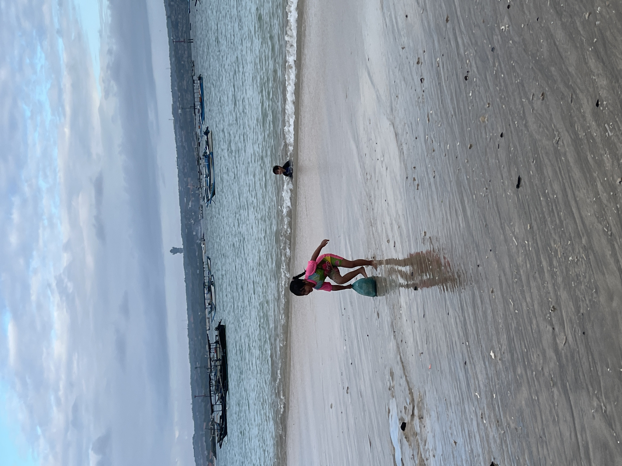 A young girl in a colorful swimsuit is playing on a wet sandy beach in Kuta, Indonesia. She is bending over, holding a green bag or bucket, and her reflection is visible in the wet sand. In the background, the ocean waves are gently rolling onto the shore, and several traditional Indonesian fishing boats are anchored in the water. Further in the distance, along the coastline, is the silhouette of a large statue against the cloudy sky, and a tall structure that appears to be a control tower or observation deck. Another child, partially submerged, is visible in the shallow water near the shore. The scene depicts a typical day at the beach with children enjoying the water and sand.