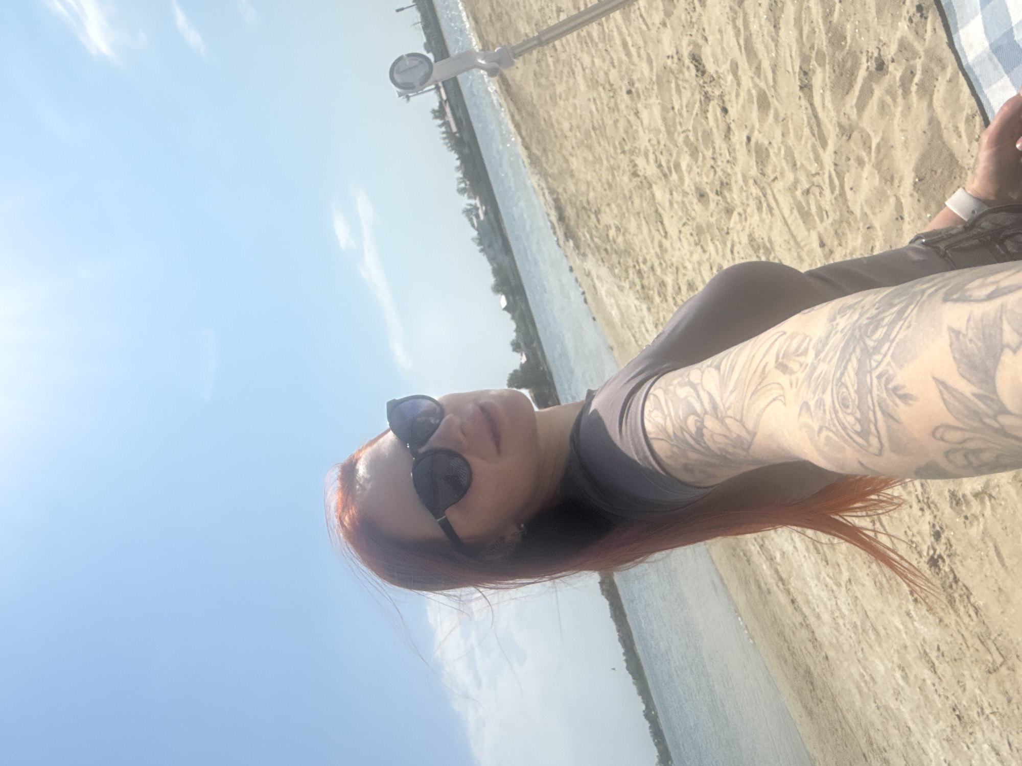 The image shows a woman with red hair and sunglasses lying on a sandy beach. She is wearing a dark-colored shirt and has extensive tattoos on her right arm. The beach is adjacent to a body of water, possibly a lake or bay, with trees and buildings visible in the distance. A tall, white pole with a gauge is also visible in the background. The location is Unknown, Unknown.