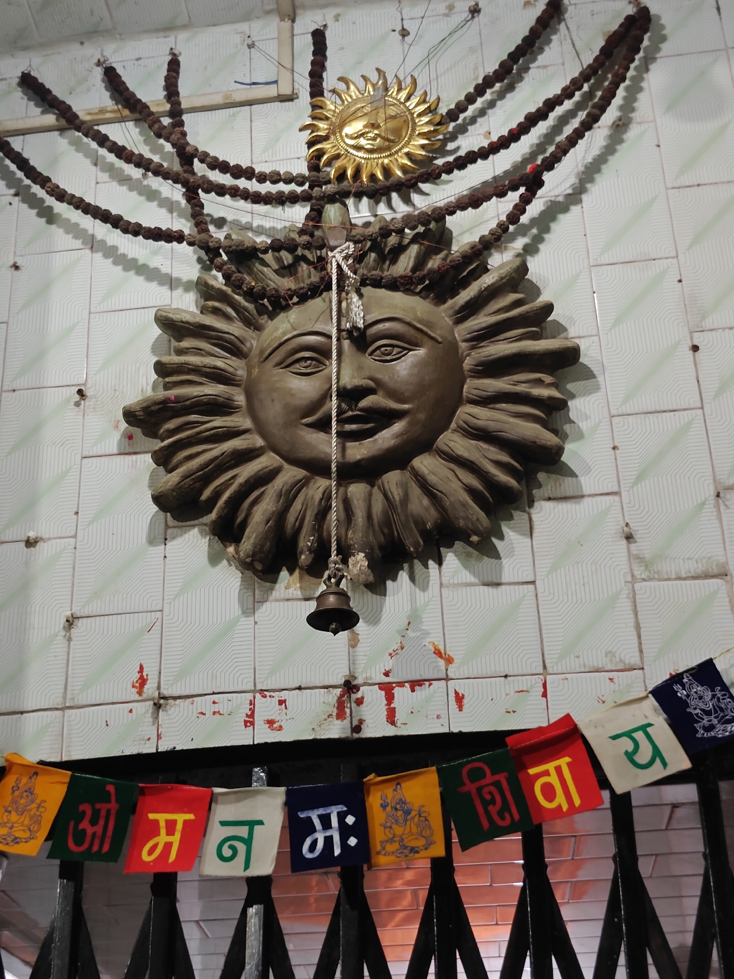 This vertical shot captures a traditional devotional display on a tiled wall, characteristic of a spiritual space in Kasol, India. At the center, a large, relief sculpture of a smiling sun deity with a benevolent, stylized face and radiating rays dominates the scene. Above this main sculpture, a smaller, golden sun motif with sharper rays is also present. Both sun effigies are adorned with multiple strands of dark, textured beads, likely rudraksha, draped in garlands from above and around them. A small brass bell hangs from a rope directly in front of the larger sun's face.
The wall itself is covered in light green and white square tiles featuring a subtle geometric pattern. It shows clear signs of age and wear, with visible dirt, missing tile fragments, and red marks, possibly from religious offerings like kumkum or vermillion.
Across the lower part of the frame, strung above a dark metal railing or gate, is a string of colorful fabric prayer flags. These flags bear inscriptions in Hindi (Devanagari script). The visible text clearly spells out parts of the sacred Hindu mantra "Om Namah Shivay" (ओम् नमः शिवाय), dedicated to Lord Shiva. Specifically, "ओम्" (Om), "न" (na), "मः" (mah), "शि" (shi), "वा" (va), and "य" (y) are discernible on separate flags. Some flags also feature white outlines of Hindu deities.
The scene suggests an indoor or covered space, illuminated by artificial light, contributing to a calm and spiritual atmosphere. No direct activity or people are shown, but the arrangement of religious artifacts implies an active place of worship or reverence.