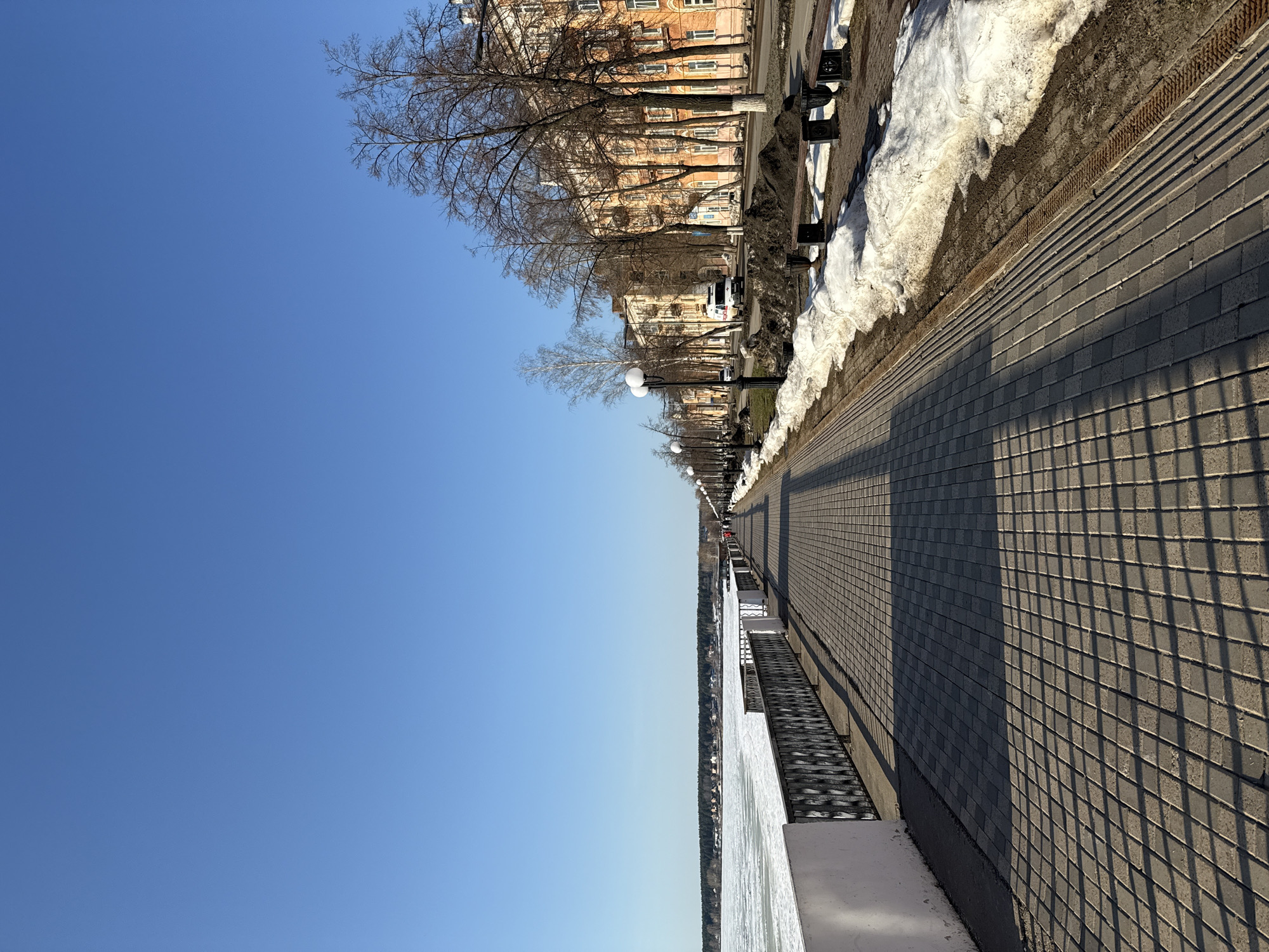 The image shows a paved walkway along a riverfront in Votkinsk, Russia. The walkway is made of gray, rectangular paving stones and runs along a white concrete barrier overlooking the frozen river. On the other side of the walkway is a row of buildings with orange facades. Trees line the street between the buildings and the walkway. Patches of snow remain on the ground near the buildings and benches. The sky is clear and blue. The image is taken at an angle, emphasizing the length of the walkway and the perspective.