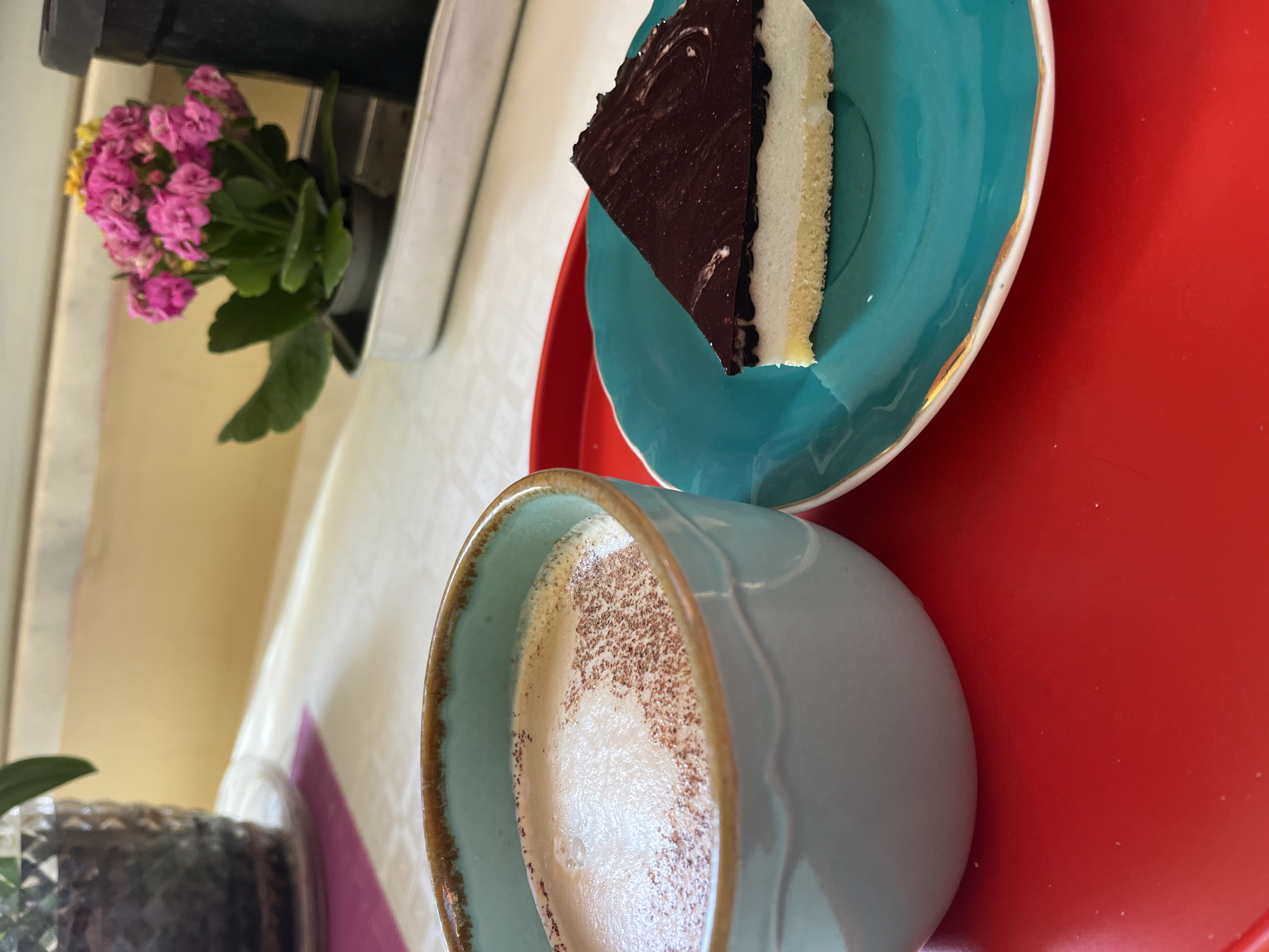 The image presents a close-up, high-angle view of a table setting. A light blue ceramic mug, partially filled with a foamy beverage sprinkled with brown powder (likely cocoa or cinnamon), sits prominently in the foreground. Behind the mug, on a red tray, a slice of cake rests on a turquoise plate with a gold rim. The cake appears to be a layered dessert, with a dark chocolate top and a lighter-colored filling. To the left of the setting, out-of-focus, are elements that suggest a restaurant or cafe setting: a pot with pink flowers, and glimpses of table surfaces and decorative elements. While not explicitly stated, the prompt implies that this image originates from Alanya, Turkey.