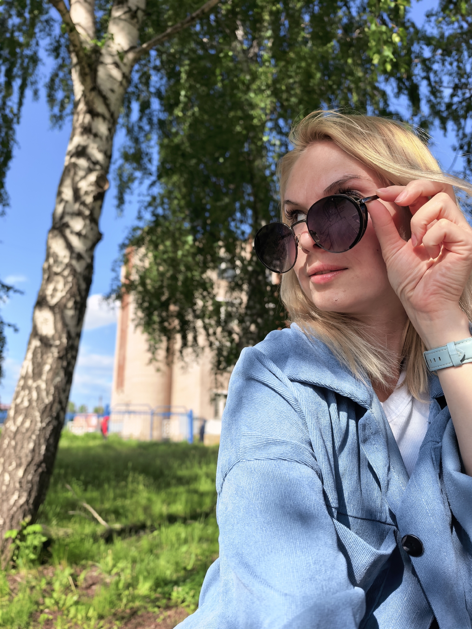 A blonde female, wearing a light blue jacket over a white t-shirt, is depicted outdoors in Sterlitamak, Russia. Her left hand is raised, adjusting dark, round sunglasses on her face, revealing a portion of her left eye and brow as she looks towards the left of the frame. A light blue wristwatch is visible on her left wrist. To the left, a birch tree with distinctive white and black bark occupies the foreground. The background consists of green grass, blue metal fencing, and a large, out-of-focus brick building, consistent with an industrial structure. The sky is clear blue with minimal cloud cover, and the scene is illuminated by strong daylight.