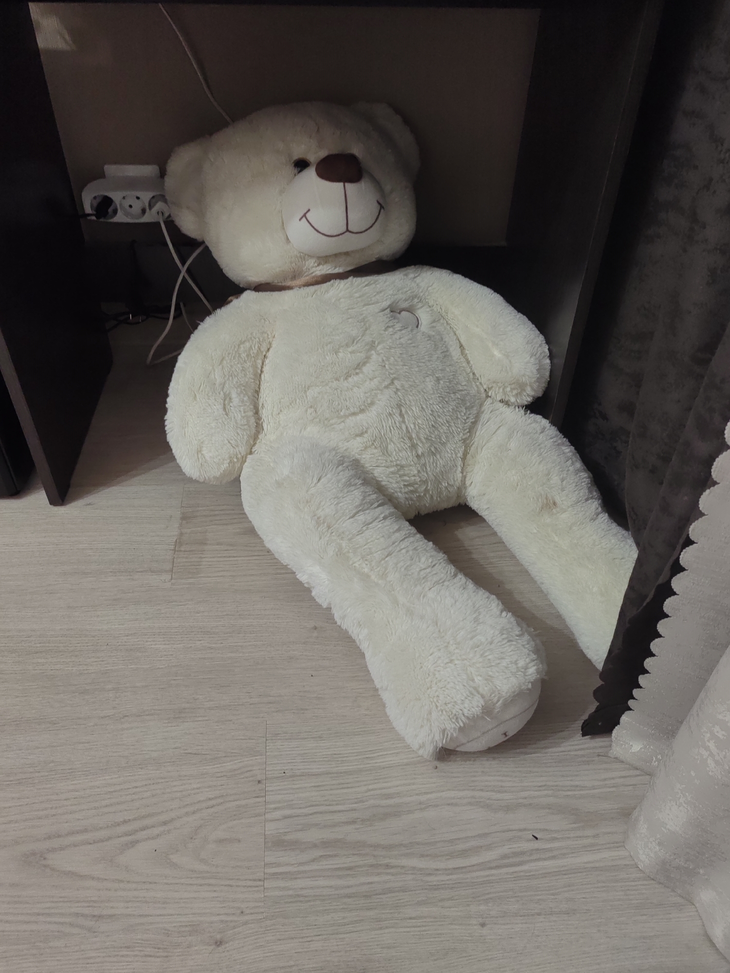 A large, white, plush teddy bear is lying on a light-colored, wood-planked floor, partially beneath a dark brown piece of furniture. The teddy bear is positioned on its back, with its legs splayed out. Its head is turned slightly to the left, revealing its smiling face with a brown nose and stitched mouth. A thin, dark brown ribbon is tied around its neck. In the background, a white multi-outlet power strip with two electrical cords is visible, plugged into the wall or furniture. To the right of the teddy bear, a dark brown curtain and a white, ruffled fabric are partially visible. The scene appears to be indoors, likely in a residential setting. The lighting is dim, casting shadows. The image does not contain any information that would allow it to be located in the city of Ostafievo, Russia.