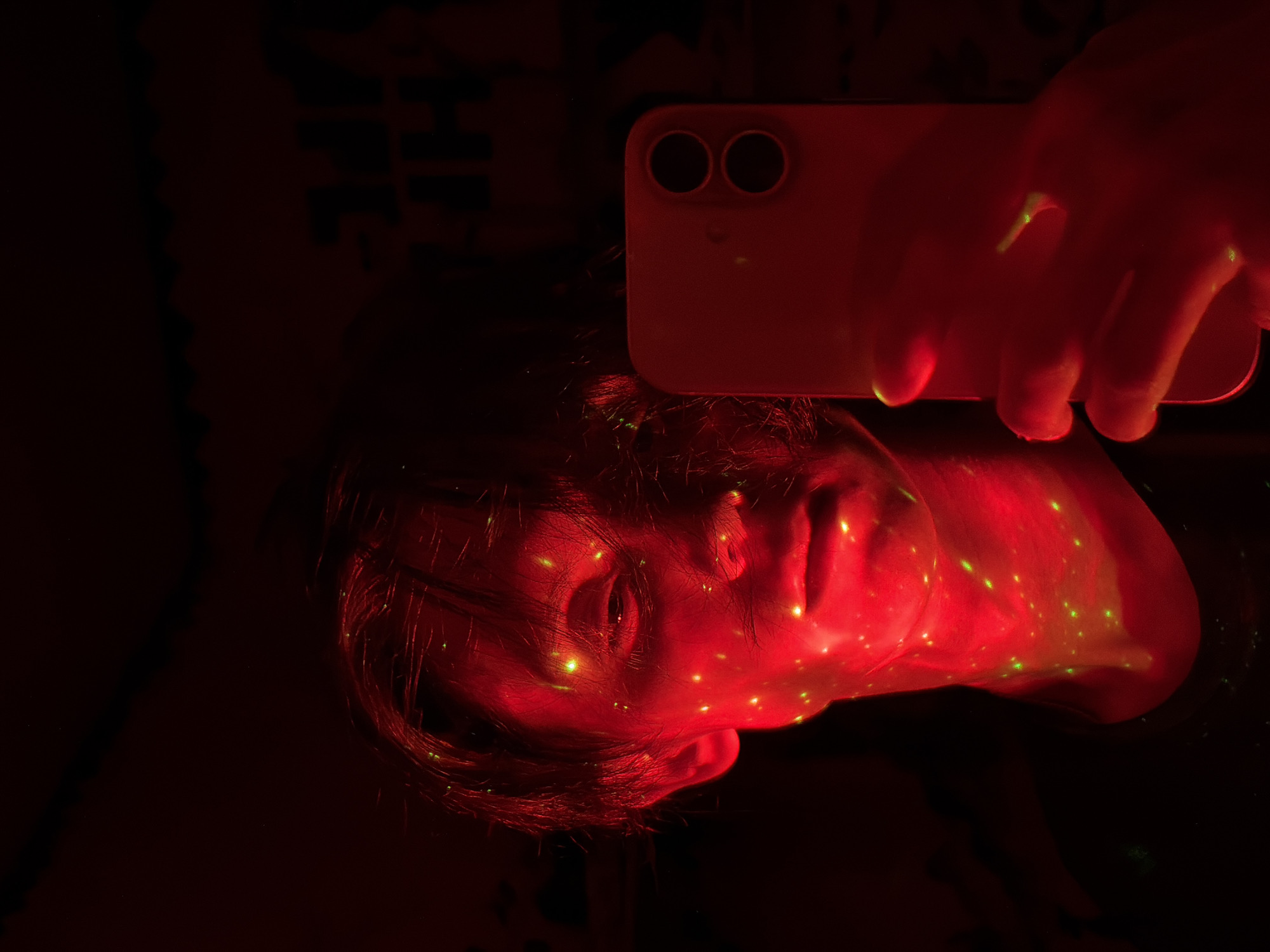 An image depicts a person holding a smartphone in their right hand, taking a selfie. The scene is illuminated by red light, with green speckles scattered across the person's face and neck. The person's left eye is visible and appears to be looking at the phone's screen. The background is dark and indistinct, with some faint, blurry shapes that might be text. The smartphone is a red iPhone, with its rear camera lenses facing the viewer. The image is underexposed, making it difficult to discern fine details.