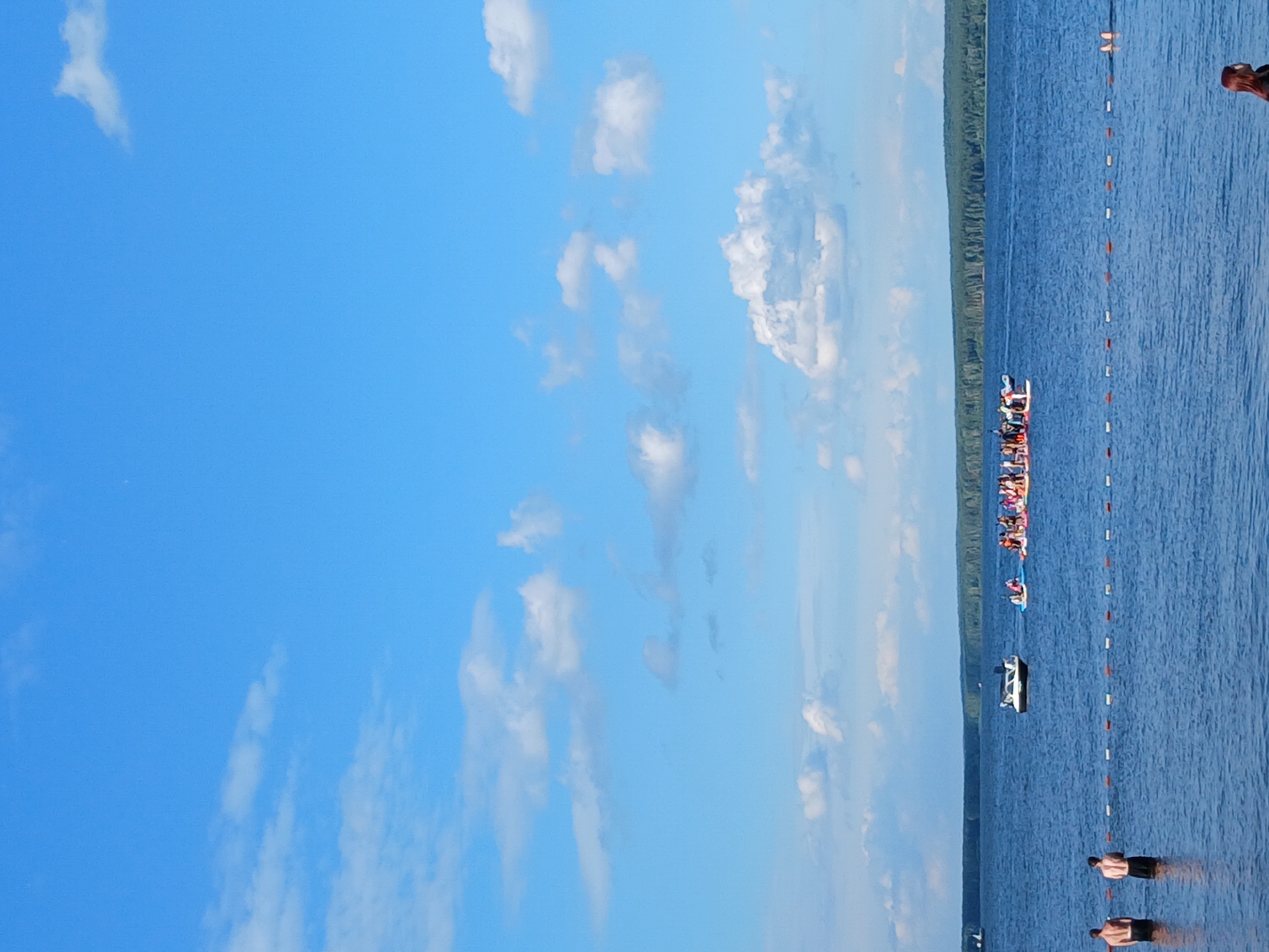 This is a photograph taken from a body of water, likely a lake or large river, under a clear blue sky with scattered white clouds. In the foreground, two men stand in the water, facing away from the camera. A rope with red and white buoys marks a boundary in the water. In the middle ground, a white motorboat is visible, and further to the right, a group of people are gathered on several paddleboards. In the background, a dense line of green trees forms the shoreline. The location is identified as Chaikovsky, Russia. The overall scene depicts a summer day at a recreational water area.