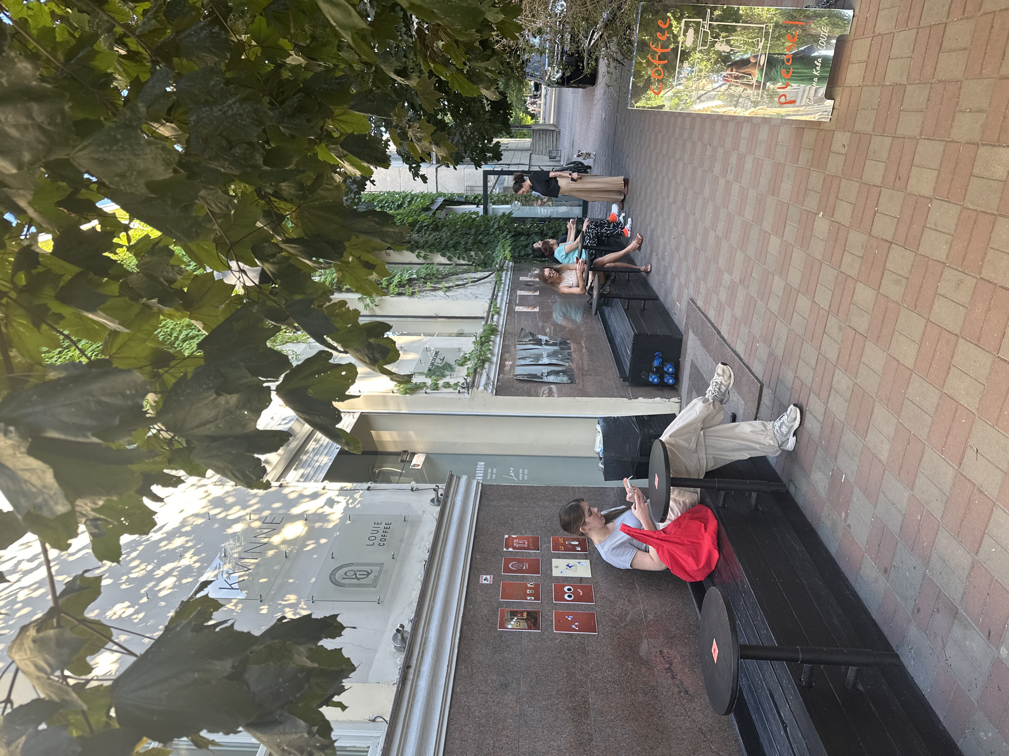 The image shows a street scene in Odesa, Ukraine, with several people sitting on a bench outside a building. The time of day appears to be afternoon, with dappled sunlight filtering through the leaves of a large tree in the foreground. The weather seems pleasant, with people dressed in casual summer clothing.
The main subjects are several individuals, including a woman in the foreground sitting on a long black bench and looking at her phone. She is wearing a light blue t-shirt, light-colored pants, and white sneakers. A bright red bag is on the bench next to her. Behind her, other people are also seated on the bench, some looking at their phones or engaged in conversation.
The setting is an urban environment, likely a street or pedestrian area in front of a commercial establishment. The building in the background has a modern facade with large windows and ivy climbing up its walls. A sign with the text "LOUIE COFFEE" is visible on the building. Adjacent to the bench, there is a sidewalk paved with red and grey tiles. Further down the street, a reflective sign displays the word "coffee" in orange lettering and what appears to be a menu or advertisement. In the distance, a small white dog can be seen on the sidewalk.
Several small framed pictures or posters are displayed on the wall of the building, some featuring cartoonish faces. The overall atmosphere suggests a relaxed, casual scene of people enjoying a break outdoors.