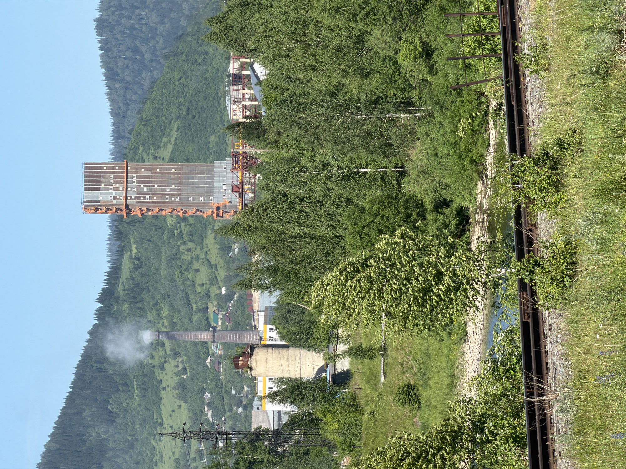 The image depicts an industrial scene set against a backdrop of lush green hills and a clear blue sky, likely in Tashtagol, Russia. In the foreground, a set of railroad tracks runs parallel to a body of water, bordered by verdant grass and foliage.
Prominently featured is a tall, unfinished industrial structure, its metallic frame and concrete sections suggestive of ongoing construction or a long-term project. Adjacent to this, a classic brick smokestack emits a plume of white smoke, indicating active industrial operations. Several buildings, likely part of the industrial complex or a nearby settlement, are visible, some with light-colored facades and yellow accents.
The overall impression is one of an active industrial site integrated into a natural landscape. The bright sunlight suggests it is daytime, and the clear sky indicates fair weather. There are no visible people in the frame, and no discernable text. The scene evokes a sense of industrial activity within a natural, possibly remote, environment.