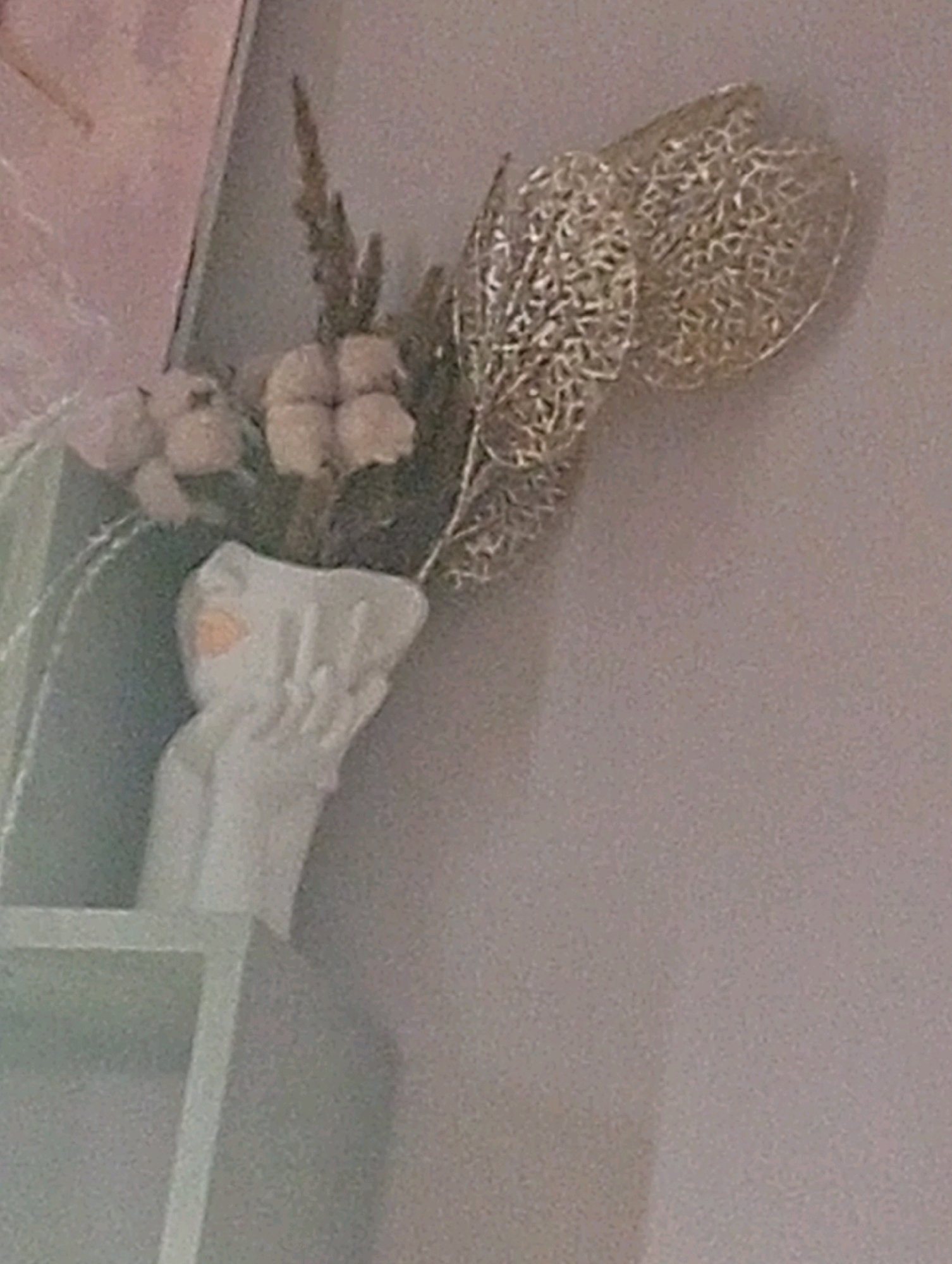 This is a close-up, blurry, indoor photograph of a decorative arrangement. The arrangement includes a small white ceramic vase with an abstract, sculpted design, possibly resembling hands or fabric. Inside the vase are dried flowers or artificial floral elements, including what appears to be cotton bolls and a feathery spike. A delicate, metallic filigree ornament, possibly gold or brass, is also part of the arrangement, extending upwards and to the right. The background is a plain, light-colored wall. The foreground shows a sliver of a light-colored shelf. There is no indication of the city of Lesnoy, Russia, or any forensic details pertinent to a crime scene.
