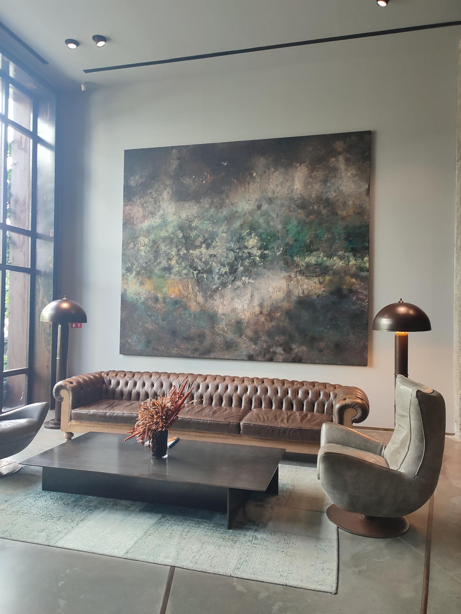 The image depicts a modern living room setting, likely within a hotel or a high-end residential space in Sarıyer, Turkey. The room features a large abstract painting on a light gray wall, dominated by dark, earthy tones with streaks of green and yellow, suggesting a landscape or atmospheric effect. In front of the painting is a long, tufted brown leather Chesterfield sofa. A dark, rectangular coffee table sits before the sofa, adorned with a vase of dried, reddish-brown flowers and a magazine. To the right of the sofa, a modern, gray swivel armchair with a circular base is positioned. Two industrial-style floor lamps with dome-shaped shades are present, one to the left of the sofa and another to the right, both casting a warm light. A large window with dark mullions is visible on the left side of the frame, allowing natural light to enter. The floor appears to be polished concrete, with a light-colored, patterned rug beneath the coffee table and sofa. The overall ambiance is one of sophisticated comfort and contemporary design, characteristic of upscale urban establishments.