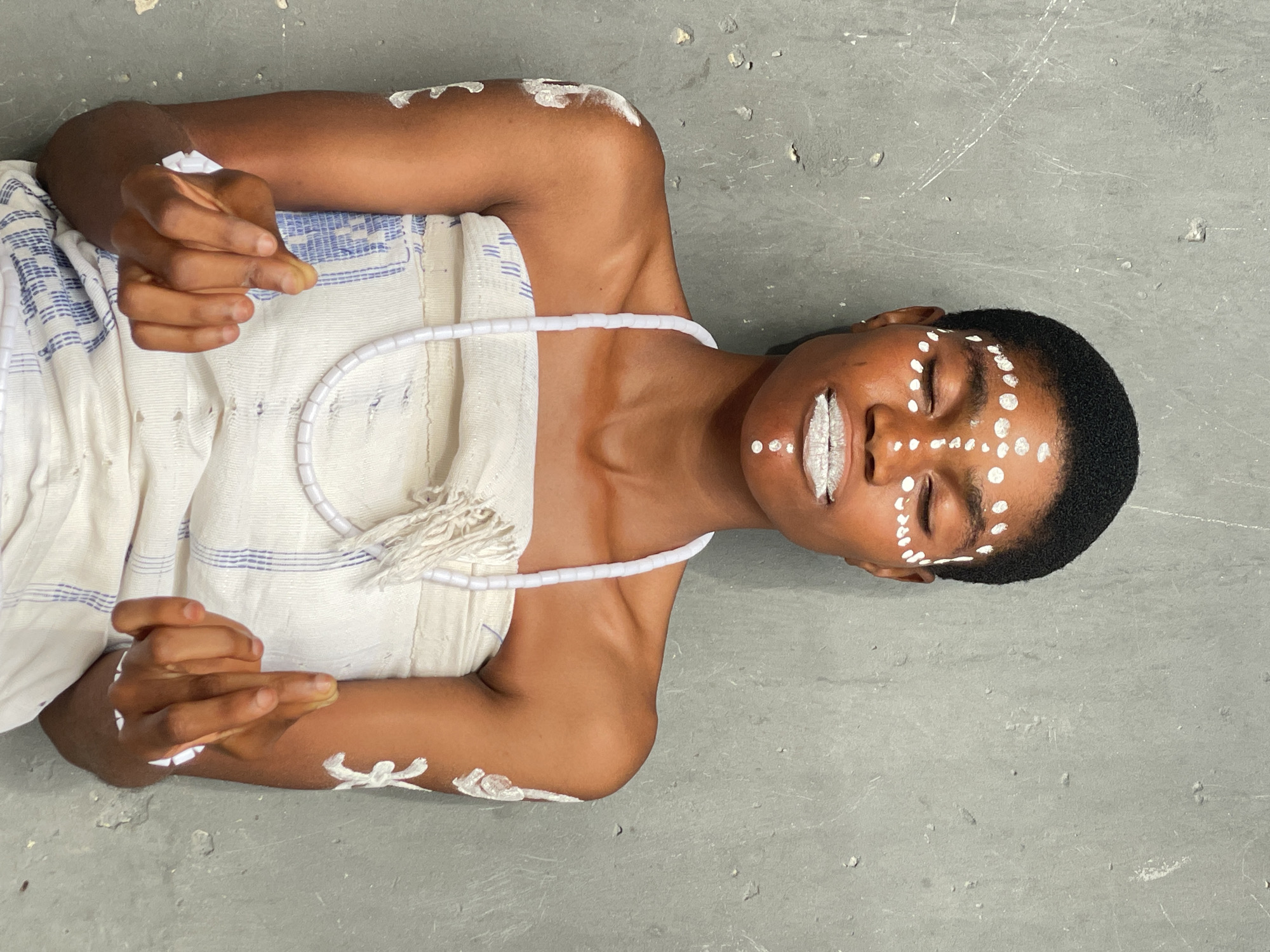 A person lies on a gray concrete surface. The person is dark-skinned and has short, dark hair. White paint is applied in patterns on the person's face, arms, and shoulders. The person is draped in a white cloth with blue stripes and wears a white beaded necklace. The person's eyes are closed, and the lips are painted white. The fingers of both hands are slightly bent. The location is likely a studio or indoor setting. There are no visible signs indicating the city of Badagry, Nigeria.