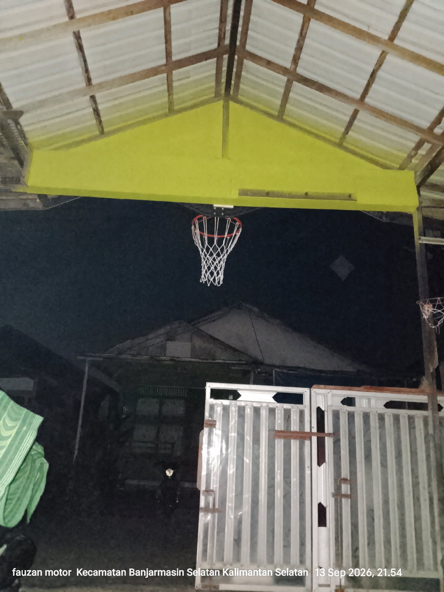 An outdoor, low-angle shot, taken at night, captures a basketball hoop hanging from a yellow-painted beam under a tin roof supported by wooden beams. The background is dark, with the silhouette of buildings and another smaller basketball hoop visible. In the foreground, a large, corrugated metal gate with vertical panels dominates the lower right portion of the image. A green, striped fabric is draped over an object on the left. Text at the bottom reads "fauzan motor Kecamatan Banjarmasin Selatan Kalimantan Selatan 13 Sep 2026, 21:54," indicating the location is in the South Banjarmasin District, South Kalimantan, Indonesia, and the date is September 13, 2026, at 9:54 PM. A motorcycle is dimly visible in the mid-ground, between the gate and the silhouetted buildings.
