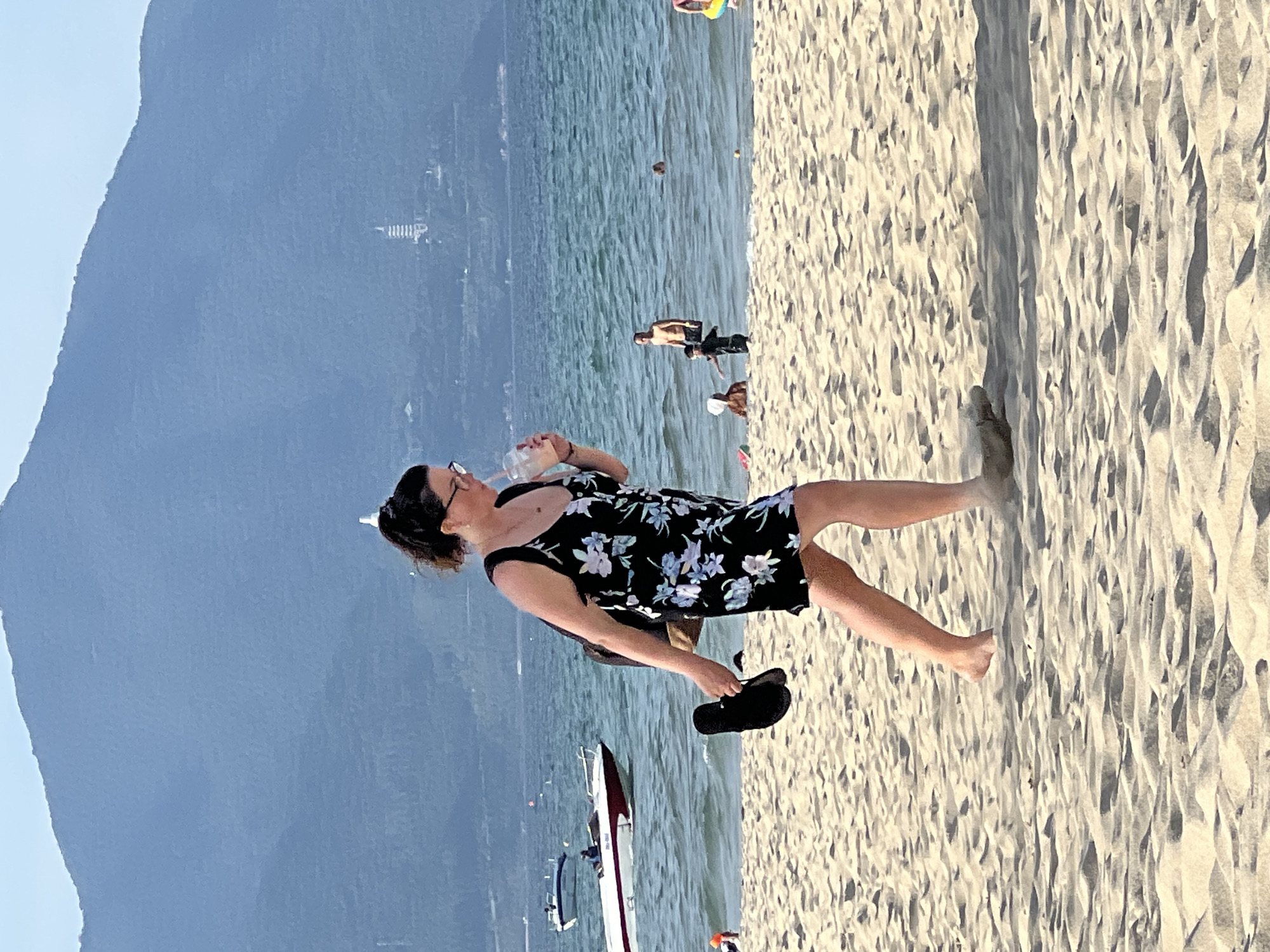 A woman with dark hair pulled back, wearing glasses, a floral print dress, and carrying a dark backpack, walks on a sandy beach. She is holding a clear plastic cup with a straw in her right hand and a black flip-flop in her left hand. The woman is walking towards the right side of the frame. In the background, the ocean is visible with several boats on the water and some people wading in the shallows. A tall, thin pagoda-like structure is visible on the mountainside in the far background. This scene is on a beach in Đà Nẵng, Vietnam, likely My Khe Beach given the typical tourist activity and mountain backdrop.