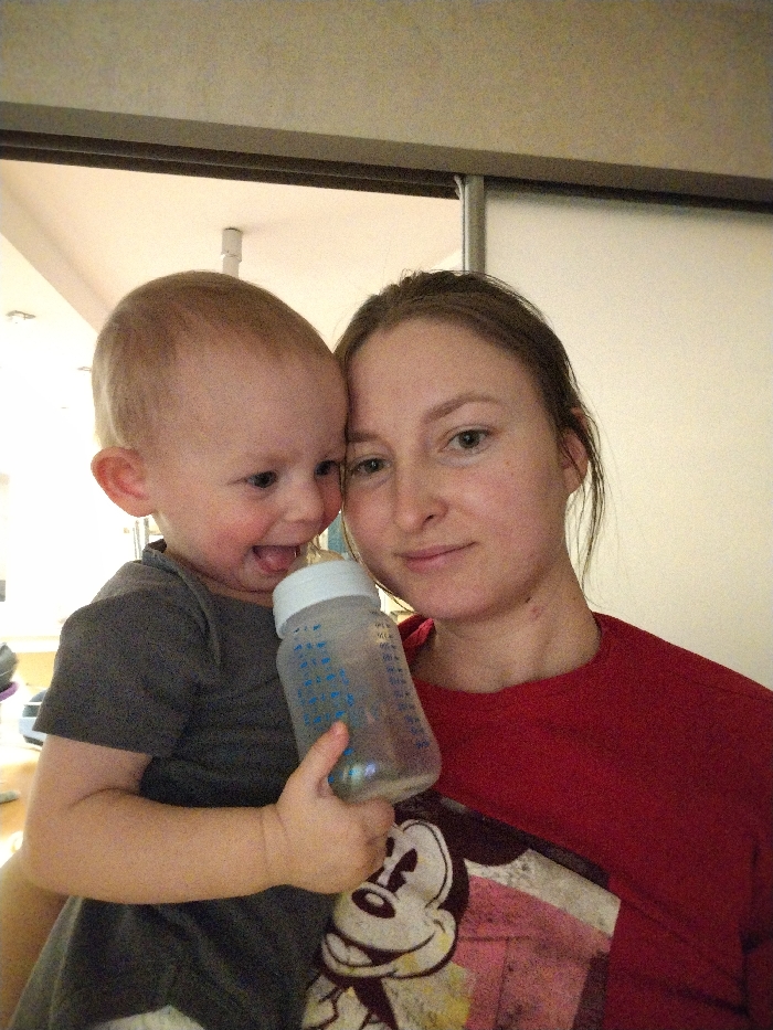 The image captures a warm, indoor portrait of a woman holding a baby or toddler.
The woman, with light skin and brown hair pulled back, looks directly at the camera with a soft, gentle expression. She is wearing a red long-sleeved top over a t-shirt that features a faded graphic of Mickey Mouse's face.
The baby, positioned on the woman's left side and slightly in front, appears to be a toddler with light-colored hair and fair skin. The child is smiling widely, with their mouth open and tongue slightly visible, looking towards the woman or just off-camera. The toddler is wearing a dark grey short-sleeved t-shirt and is holding a clear plastic baby bottle with a white cap. Blue measurement markings are visible on the side of the bottle.
The woman is embracing the child, whose head is resting against her cheek. The overall impression is one of affection and joy.
The background is an indoor setting with light-colored walls or partitions. A light fixture is visible in the upper left corner, and there are hints of a doorway or sliding panel behind them. The lighting appears soft and even, suggesting an indoor environment during the day or with artificial lighting. There is no clearly legible text other than the measurement markings on the bottle.