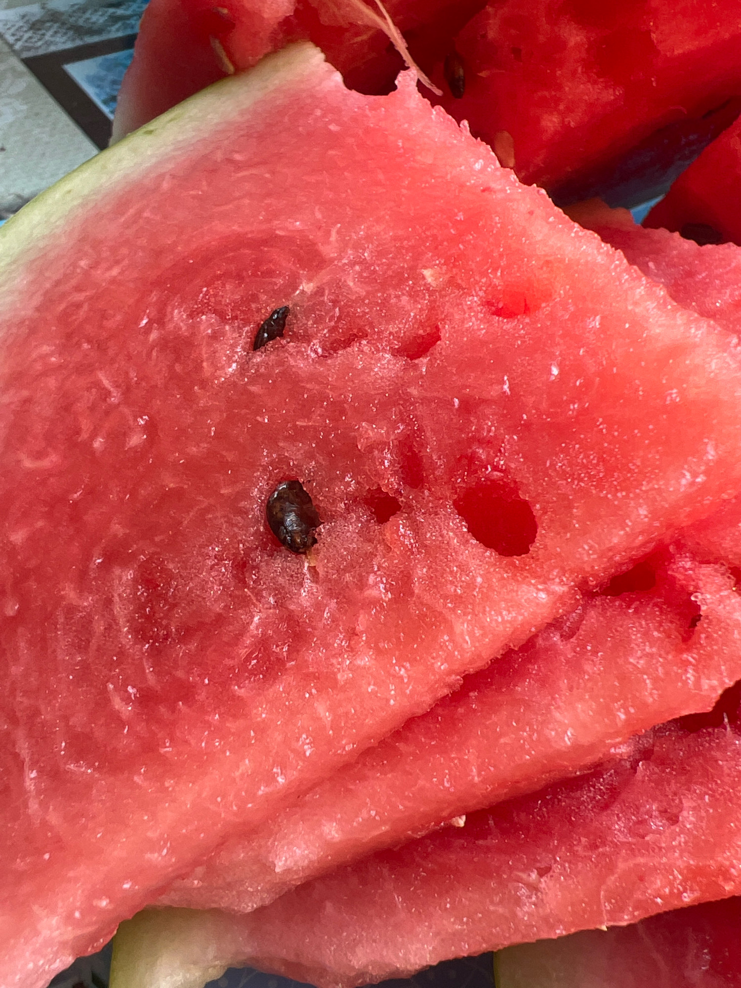 A close-up, overhead shot showcases several slices of bright red watermelon with visible seeds. The watermelon flesh is textured and appears juicy, with some areas showing a glistening quality. The slices are stacked and slightly overlapping, with the rind of one slice visible at the top left. The background is blurred, but hints of a patterned surface are discernible. The scene is indoors, with no discernible city or environmental context.