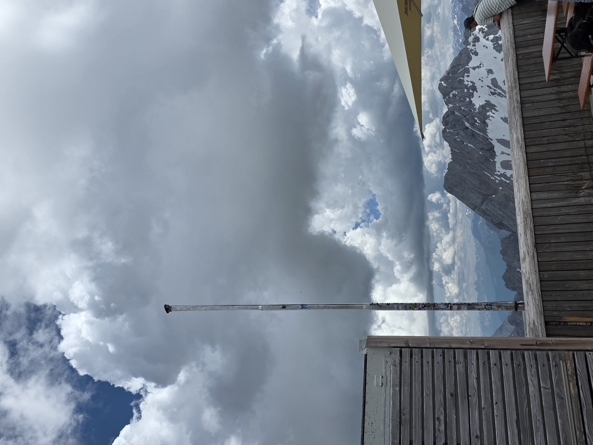 The image depicts a high-altitude viewpoint, likely at the Zugspitze, Germany's highest peak, accessible from Garmisch-Partenkirchen. In the foreground, a wooden railing separates a viewing platform from the mountain landscape. To the left, a section of a building with horizontal wooden siding is visible. A tall, slender metal pole, adorned with numerous stickers, stands vertically near the center of the frame, extending towards a dramatic, cloudy sky. Below the pole, a wooden deck with benches and tables suggests an area for visitors. A person, wearing a striped shirt and a cap, leans over the railing, looking out at the majestic, snow-capped mountains in the distance. The sky is filled with a mix of dark, imposing storm clouds and patches of bright blue, indicating dynamic weather conditions. The overall scene conveys a sense of vastness and the raw beauty of a mountain environment.