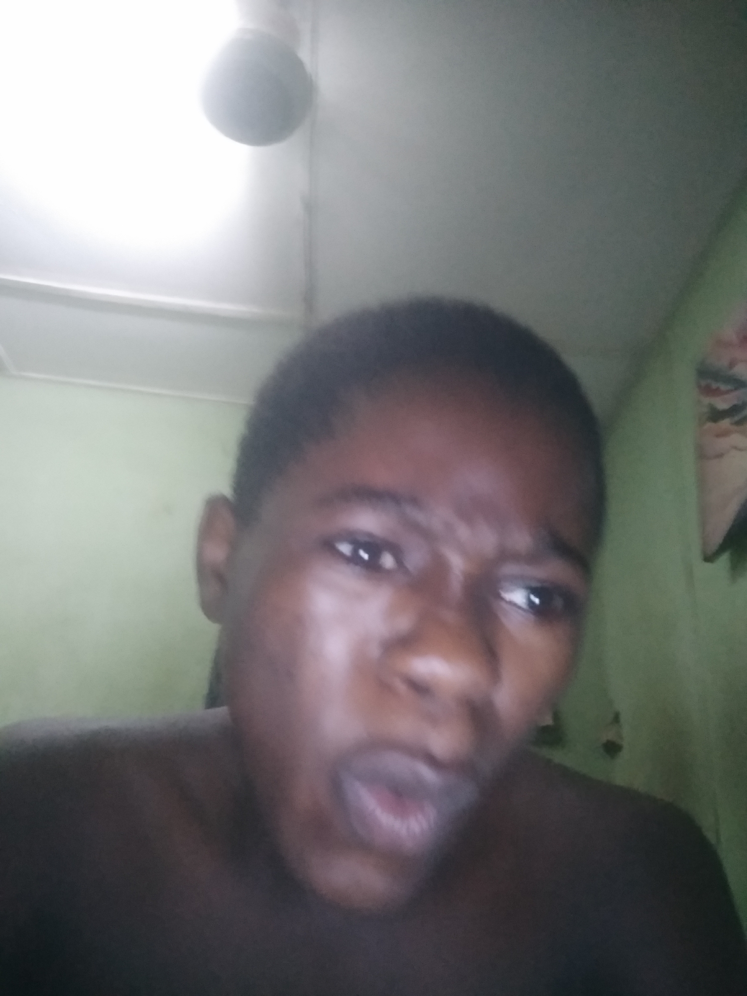 A close-up, indoor selfie shows a person with dark skin and short, dark hair. The person's facial expression includes furrowed brows and an open mouth. A light-colored ceiling fan is visible in the upper portion of the image. The background includes light green walls. The location of the image is Shangisha, Nigeria.