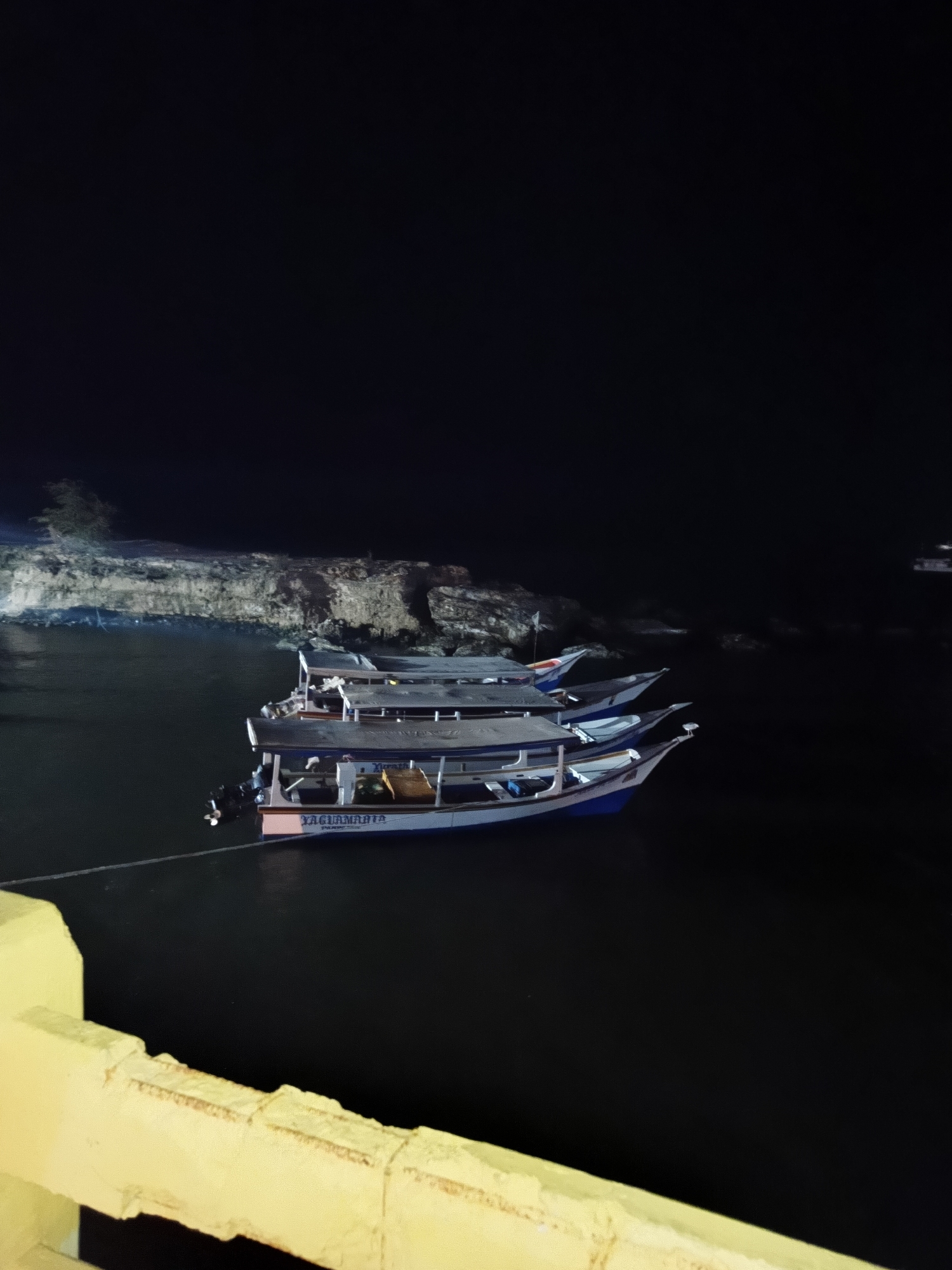 The image depicts boats docked in the bay of Juan Griego, Venezuela, at night. There are at least four boats tied together. The lead boat has "YACUAMARINA" written on its side. In the background, illuminated by lights, is a rocky shoreline. A yellow concrete structure, likely part of a walkway or bridge, is visible in the lower left corner of the frame. The water is dark and still.