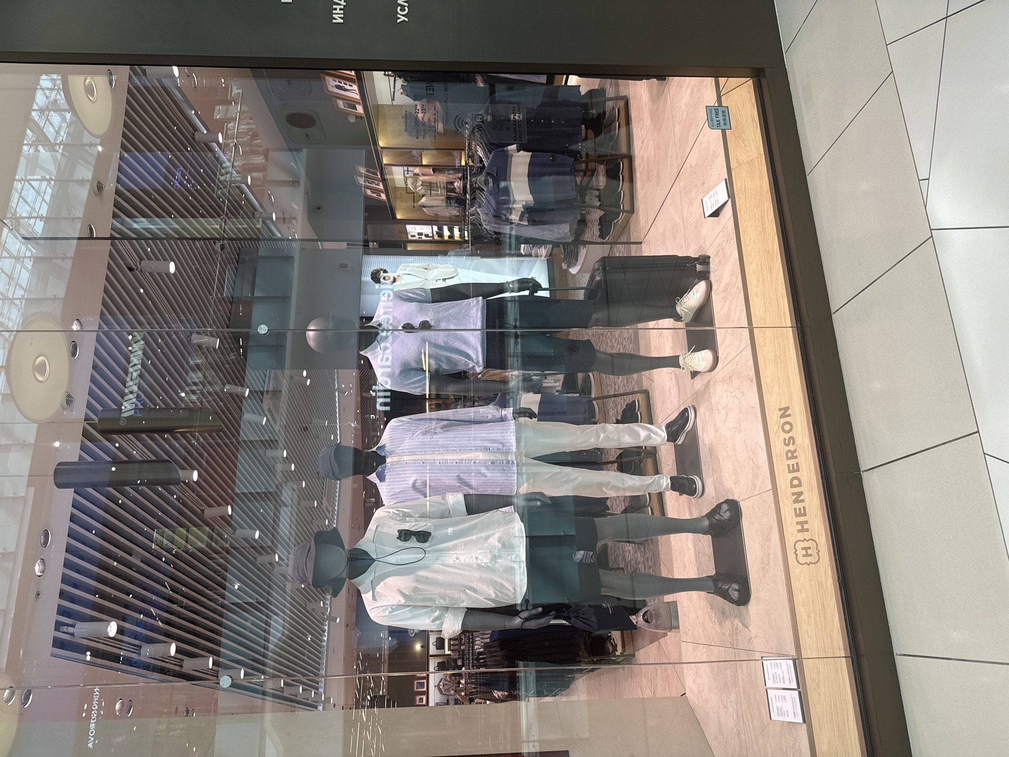 This media file captures a retail store window display, likely in a shopping mall. The primary subjects are several mannequins dressed in men's casual wear, presented in a staged display. One mannequin in the foreground wears a white collared shirt, dark shorts, and black sneakers. Another mannequin behind it is dressed in a light blue and white striped shirt with white pants and black sneakers. A third mannequin, further back, is clad in a dark blue jacket and pants. The display is set within a store interior with clothing racks and shelving visible in the background.
The setting appears to be a bright, well-lit retail environment, suggested by the overhead lighting fixtures and the polished flooring. Reflections on the glass window show the interior of the store and some elements of the surrounding mall, including what looks like another store's window with framed items and clothing racks.
The notable text present includes a store name, "H HENDERSON," prominently displayed on a wooden element of the window frame. Other visible text includes what appears to be store hours in Russian ("BCNT 1000-2200," "ПТ-СЕ 1000-2300") and "TAX FREE" with Chinese characters, indicating potential tax-free shopping services for international customers. There are also some partially visible Russian words at the top of the frame. The overall impression is a clean, modern retail display designed to attract customers. No specific time of day or weather conditions can be determined from the image.
