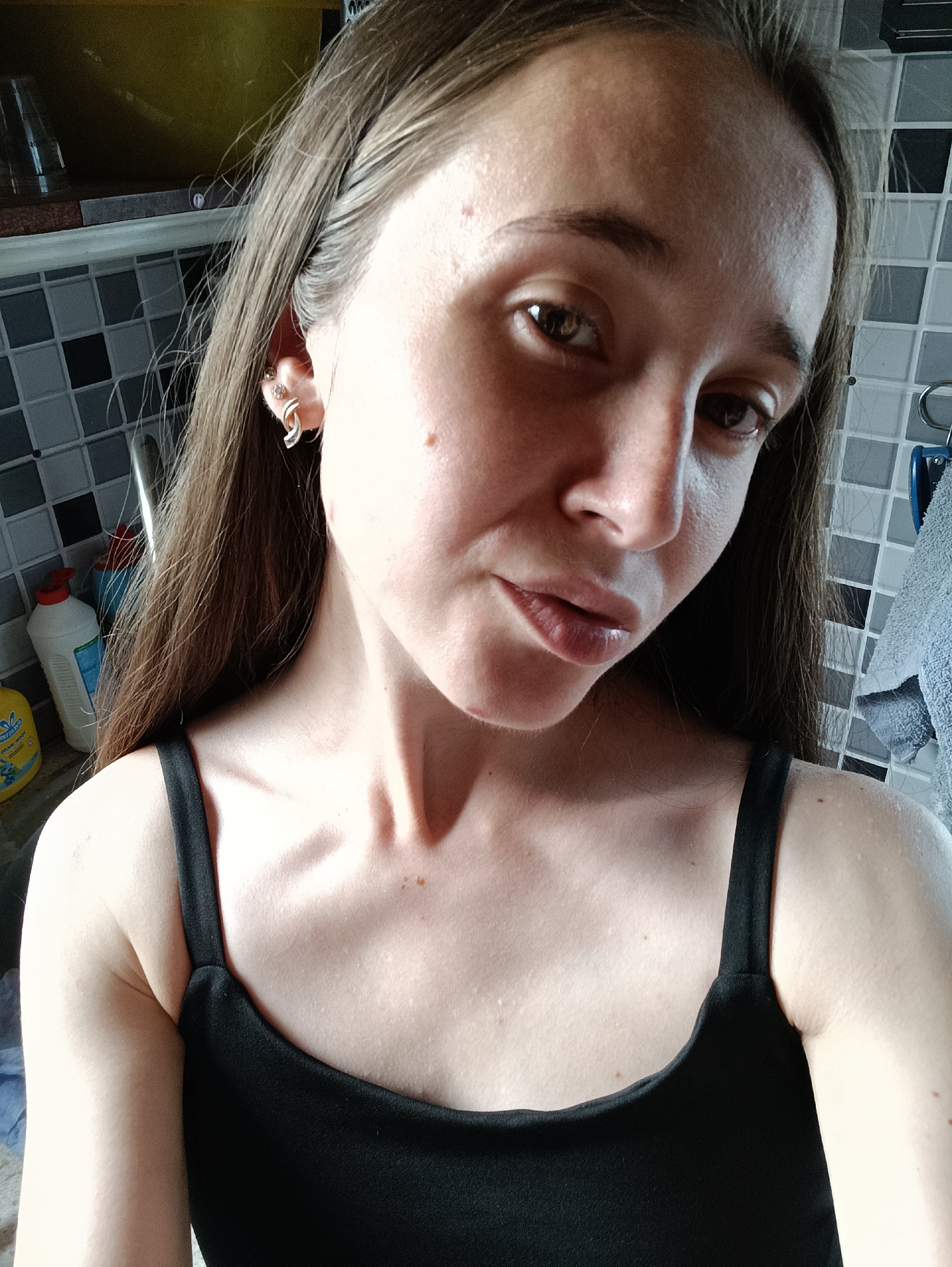 A young woman is captured in a close-up selfie, looking directly at the camera. She has long, dark brown hair, parted in the middle and falling over her shoulders. Her skin is fair with a slight tan, and she has dark brown eyes. She is wearing a black tank top, revealing her shoulders and collarbone. Her right ear is adorned with two piercings, one with a small stud and another with a silver hoop earring. The background suggests an indoor setting, possibly a kitchen, with a tiled backsplash and shelves holding bottles of cleaning products. There are no obvious signs of the city of Vypasne, Ukraine. The lighting is bright, casting some shadows on her face and chest.