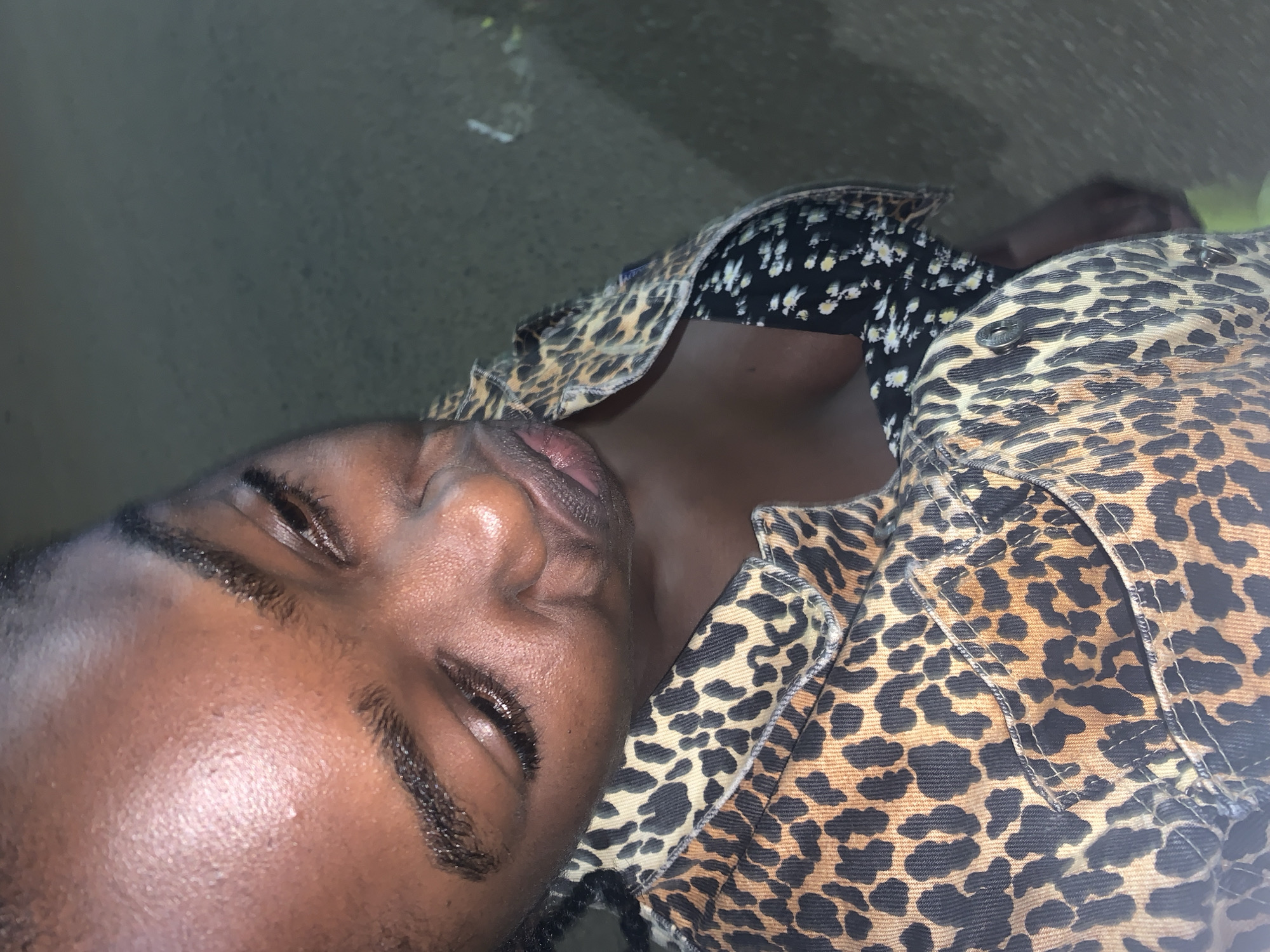 This close-up portrait, originally rotated, features an individual with dark skin, likely female, reclining or lying down and looking upwards. The person's eyes are mostly closed, with one eye slightly ajar, revealing dark eyelashes and well-defined eyebrows. Their lips are full, slightly parted, and appear moist. A subtle sheen is visible on their skin, possibly due to light reflection or moisture.
The individual is dressed in a prominent leopard-print jacket or heavy shirt, which is worn over a dark top patterned with small white flowers. Details of the leopard-print fabric, including stitching and button-like elements, are visible on the garment's collar and body. The background is dark and largely out of focus, resembling an indistinct, possibly concrete surface. A faint, blurry suggestion of a lighter, perhaps green, object can be seen in the upper right.
The lighting appears dim, suggesting the photograph was taken in the evening, at night, or in an indoor setting with low illumination. The person's pose and partially closed eyes convey a relaxed or introspective mood. The image does not contain any visible text. The provided context indicates the location as Jos, Nigeria.