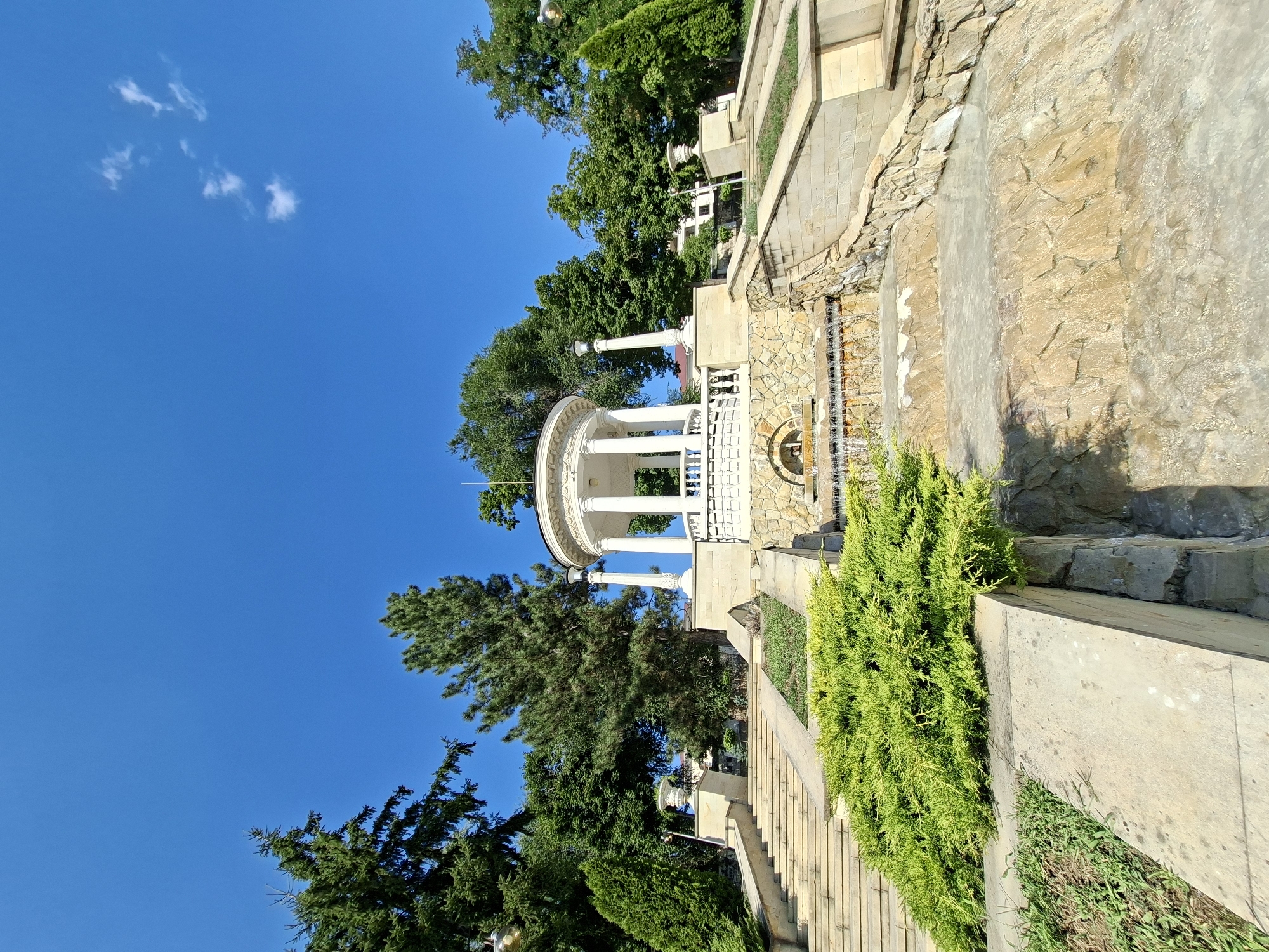 The image shows a bright, sunny day in Chișinău, Moldova, featuring the rotunda in Dendrarium Park. A white, neoclassical rotunda with Corinthian columns stands prominently, surrounded by lush green trees and a clear blue sky with a few wispy clouds. In front of the rotunda, a multi-tiered stone staircase leads down to a cascading fountain. Bright green bushes and manicured lawns complement the architecture and natural elements. The overall scene conveys a sense of tranquility and natural beauty within an urban park setting.