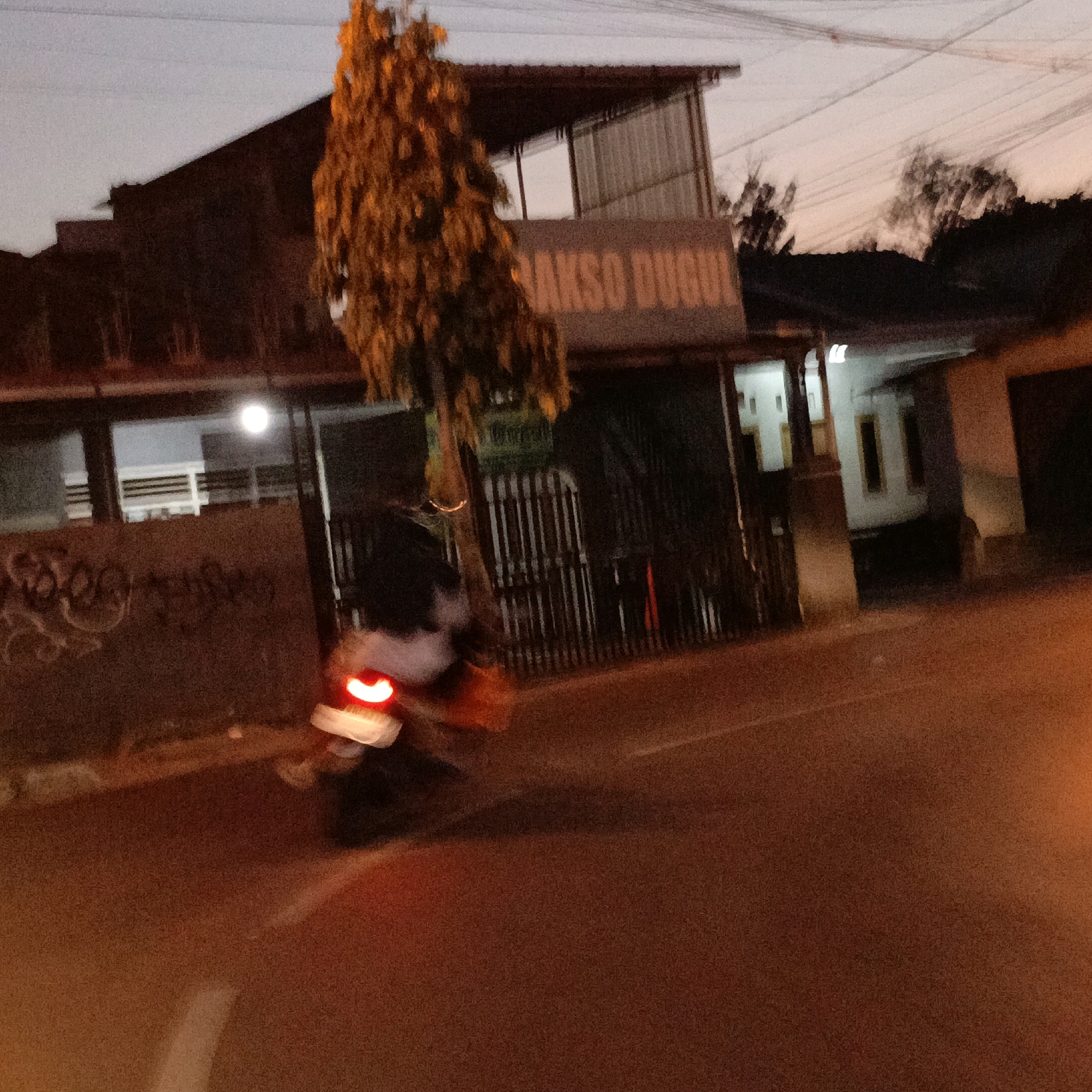 An outdoor, medium-shot, blurry image shows a street scene at dusk in Salatiga, Indonesia. In the foreground, a motorcycle with a lit red taillight is moving from left to right, its rider is indistinct. The motorcycle is on a paved road with some white road markings. Behind the road, a building with a sign that reads "BAKSO DUGUL" is visible. The building has large windows and a dark roof. To the left of the building, graffiti is visible on a wall. A tall, leafy tree with orange foliage stands in front of the building. The sky is a dusky grey.