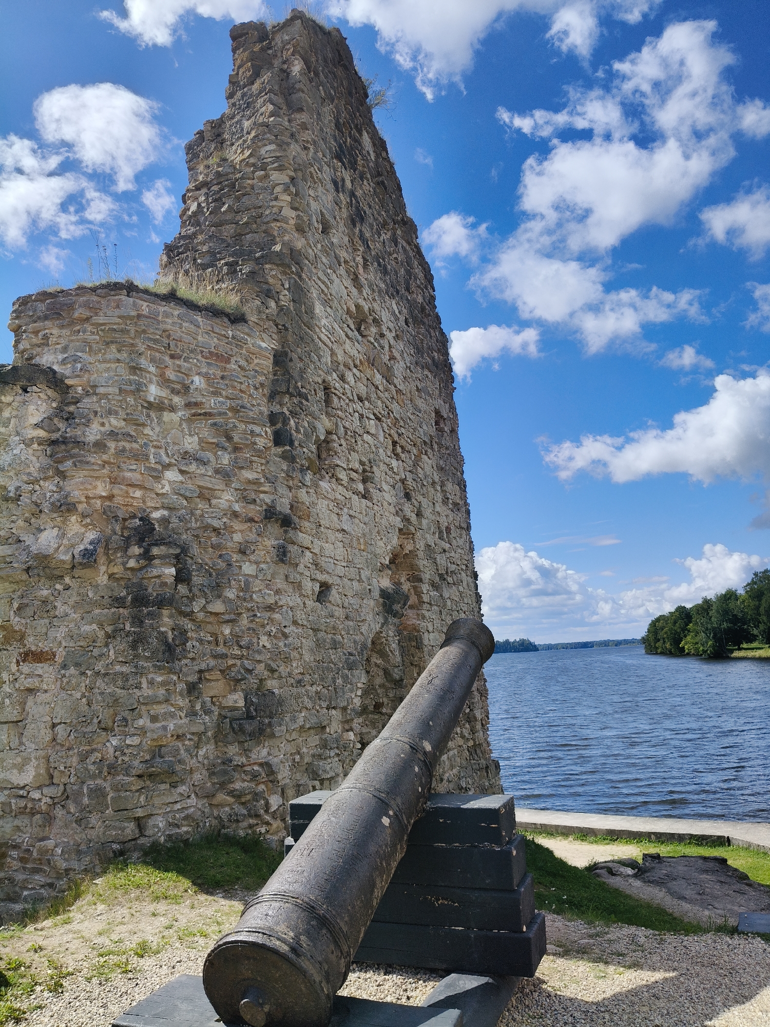 This is a photograph of the ruins of Koknese Castle in Koknese, Latvia. The image shows a large, weathered stone wall on the left, part of the castle ruins. In the foreground, a dark-colored cannon is positioned on a dark wooden stand. The cannon is pointing towards the right side of the frame. Beyond the castle ruins, a wide body of blue water, likely the Daugava River, stretches across the middle ground. The far bank of the river is lined with lush green trees. The sky is bright blue with scattered white clouds. The ground in the foreground is covered with gravel and patches of green grass. The overall scene depicts a historical site with a scenic backdrop.