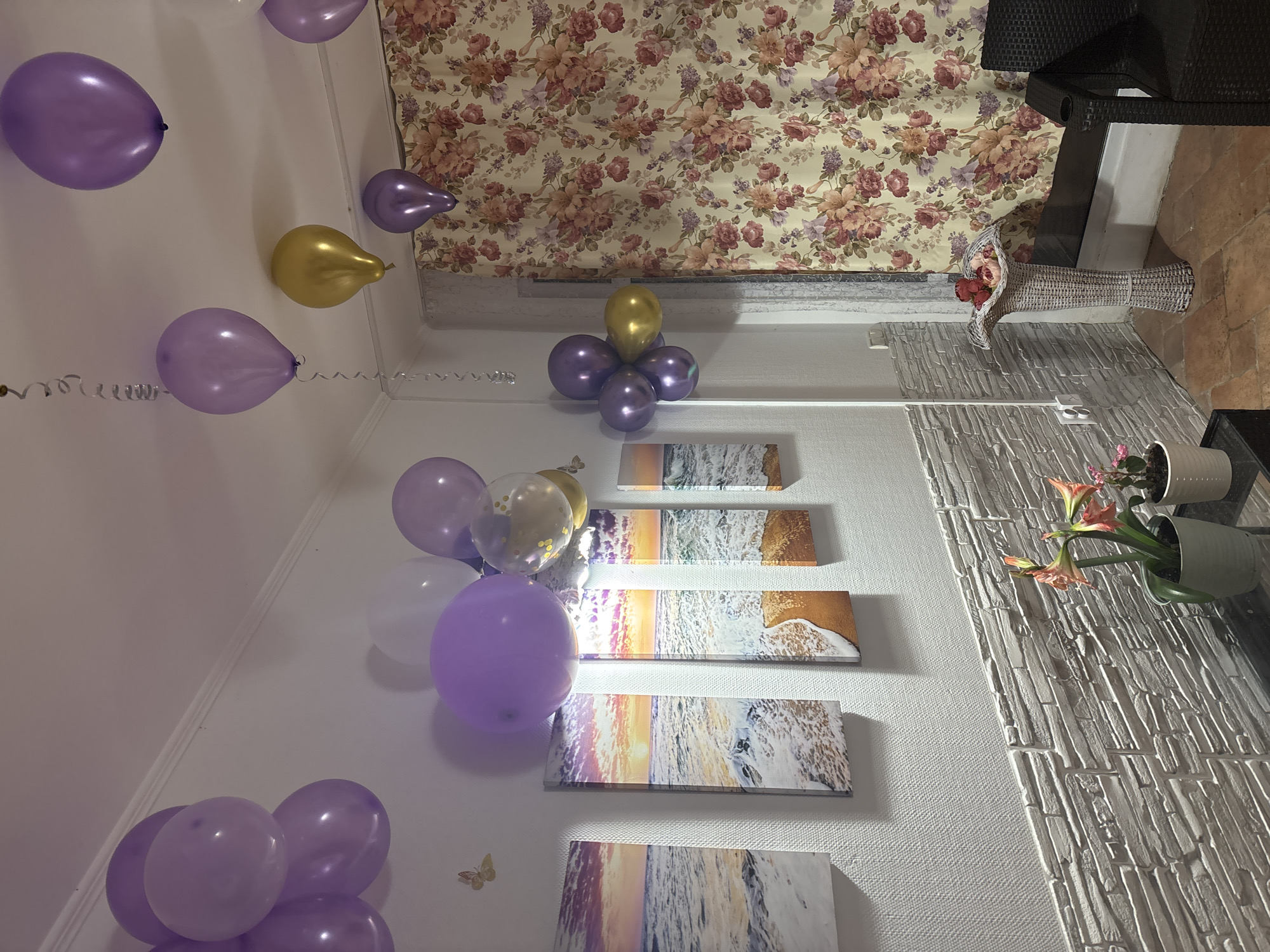 The image depicts an interior space decorated for a celebration, likely a birthday. Several purple and gold balloons are scattered throughout the room. The walls are adorned with framed artwork depicting beach scenes with sunsets and ocean waves. A decorative wallpaper with a floral pattern covers the upper portion of one wall. There is also a faux brick accent wall with potted plants. A white wicker basket with red roses is positioned near the top of the floral wallpaper. The image does not provide information about the location or the city of Sochi, Russia.
