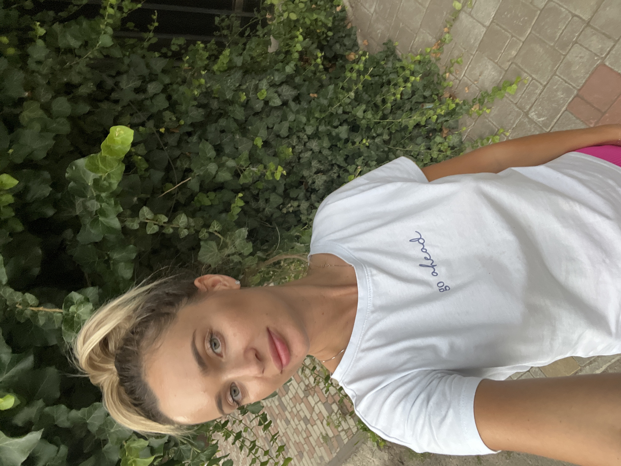 A young woman with blonde hair in a ponytail, wearing a white t-shirt with the inscription "go ahead" and pink shorts, stands in front of a wall covered in ivy. The background shows a paved path made of rectangular light-colored stones. The woman is holding a phone and taking a selfie. The setting appears to be outdoors in a residential or public area.