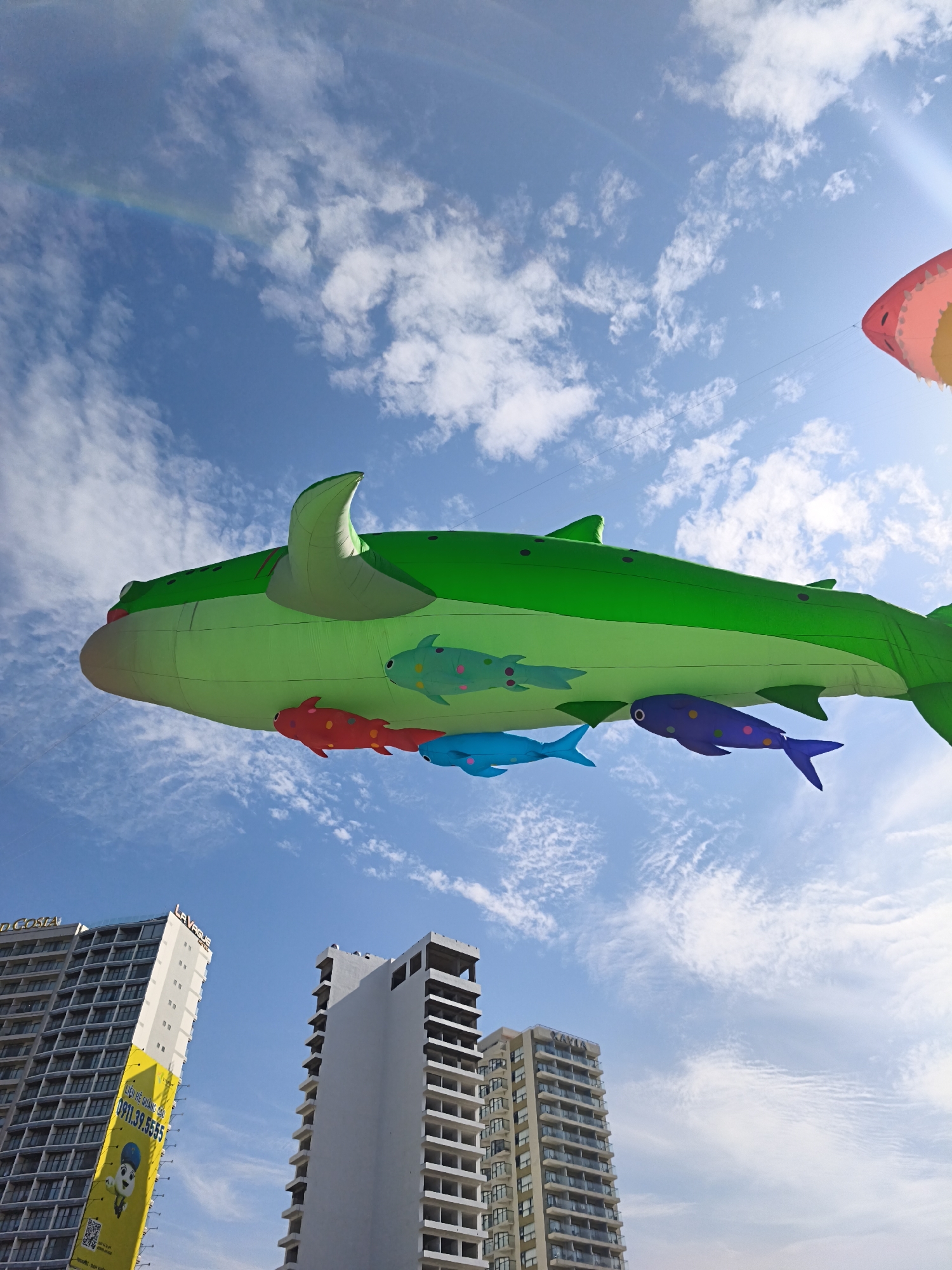 This is a low-angle outdoor photograph taken on a bright, sunny day in Nha Trang, Vietnam, with a clear blue sky dotted with white clouds. Dominating the upper portion of the frame is a large, green inflatable whale, possibly a kite, with several smaller colorful inflatable fish attached to its underside. The whale is depicted with a playful design, featuring large eyes and a fin on its head. In the far upper right corner, a portion of a pink and yellow inflatable object, possibly another kite, is visible. A faint rainbow arcs across the sky from the upper left.
In the lower portion of the image, three tall buildings are visible. The building on the left has a sign with Vietnamese text and a cartoon character, along with a phone number "0911.37.55.55". The middle building is a tall, light-colored structure with multiple floors and visible balconies. The building on the right appears to be under construction, with exposed concrete floors and no windows installed. The overall scene suggests a waterfront area or a public space where inflatable decorations are displayed against a backdrop of urban development.