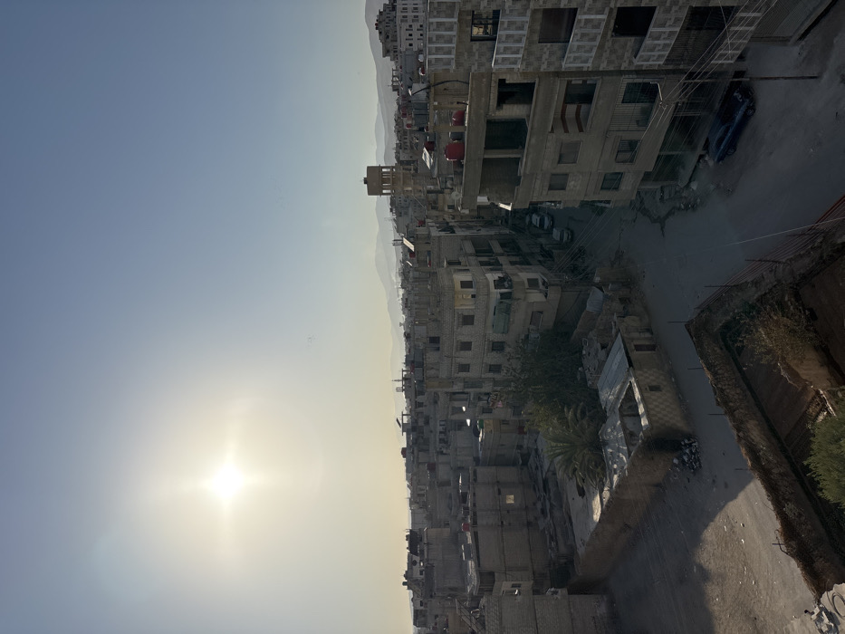 This image captures an elevated view of a residential area in Mu'adammiyat ash-Sham, Syria, under clear daylight conditions with a low sun angle. The sun is prominently featured in the upper-left quadrant, emitting a bright flare.
The scene is dominated by a dense arrangement of multi-story buildings, predominantly constructed from light-colored concrete or block materials. Many structures appear utilitarian, some exhibiting exposed rebar or unfinished sections, while others feature flat rooftops equipped with red cylindrical water tanks. Corrugated metal roofing is visible on lower annexes or damaged sections of some buildings in the foreground.
Narrow, paved and unpaved streets wind through the architectural landscape. A dark blue sedan is parked on a street in the lower-right portion of the frame. Two additional lighter-colored sedans are partially visible further down a street in the mid-ground, leading toward the center-left.
A distinctive, tall, cylindrical water tower-like structure with a square base stands in the mid-ground, slightly to the right of the image center. In the lower-middle left, a low-rise enclosed structure with a partially de-roofed or collapsed section is visible, surrounded by accumulated debris. An empty, walled, rectangular enclosure with exposed earth occupies the immediate foreground, bottom center.
Vegetation is sparse, primarily consisting of several palm trees within enclosed courtyards and scattered smaller green foliage. An intricate network of overhead power or communication lines crisscrosses between buildings and utility poles. No individuals are visible, and no overt actions or interactions are depicted. Distant, hazy mountain ranges are discernible along the horizon.
