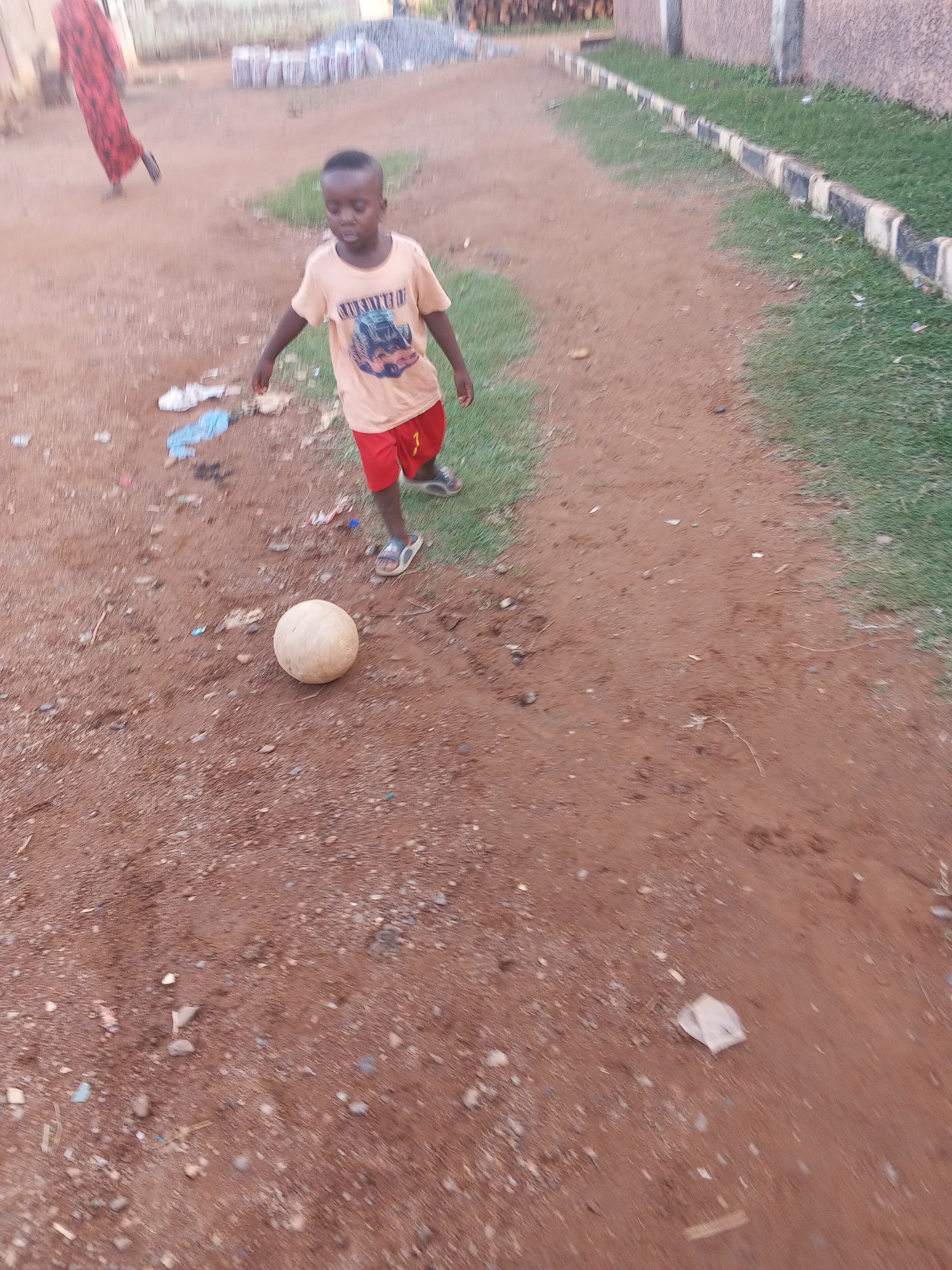 A young boy with dark hair wearing a beige t-shirt with a graphic, red shorts with a "7" on the right leg, and sandals stands on a dirt path near a white soccer ball. The path, predominantly brown dirt mixed with stones and debris, has patches of green grass on either side. A building is partially visible to the right, alongside a border of alternating black and white painted concrete blocks. Behind the boy, discarded debris, including a tattered shoe, is visible. In the distant background, wooden planks are stacked to the left of the path. A person in a red dress is walking away from the main scene. The image was likely taken in a suburban area, possibly within the city of Abuja, Nigeria.