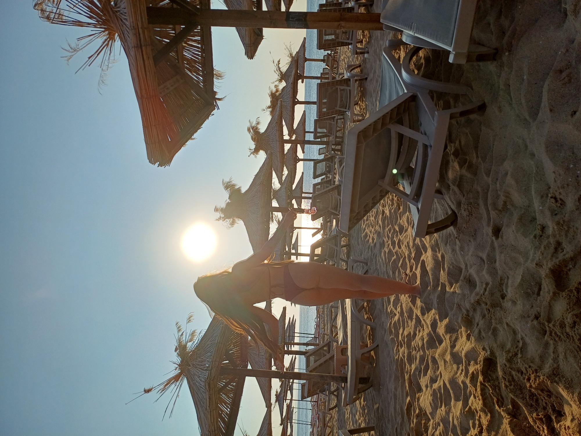 A full-length, rear view of a woman wearing a dark red bikini stands on a sandy beach. Her arms are outstretched and her long brown hair is flowing behind her. She is facing the sea, with the sun setting or rising in the background, casting a bright glare. The beach is lined with rows of empty, beige lounge chairs and straw umbrellas. The water is calm and appears to be the Black Sea. The scene is likely Varna, Bulgaria, given the characteristic straw umbrellas and lounge chairs commonly found on its beaches during the tourist season. The sand is rippled, indicating a slight breeze. The overall atmosphere is serene and suggests a peaceful vacation setting.